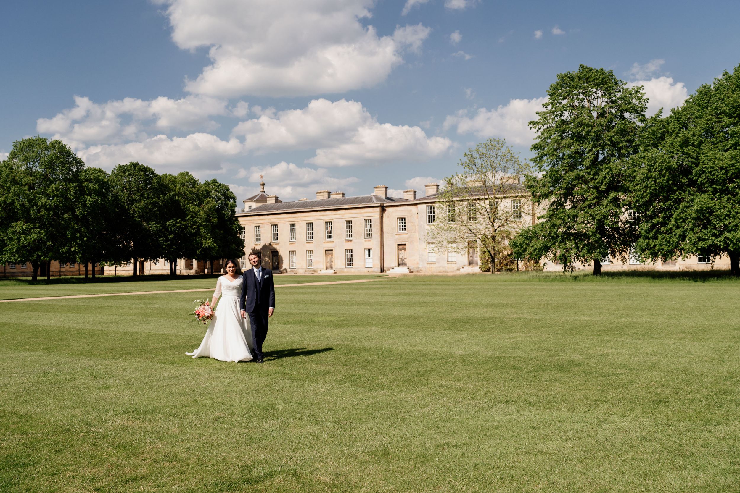 Newlyweds walking on vast green lawn in front of a historic manor house of Downing College.