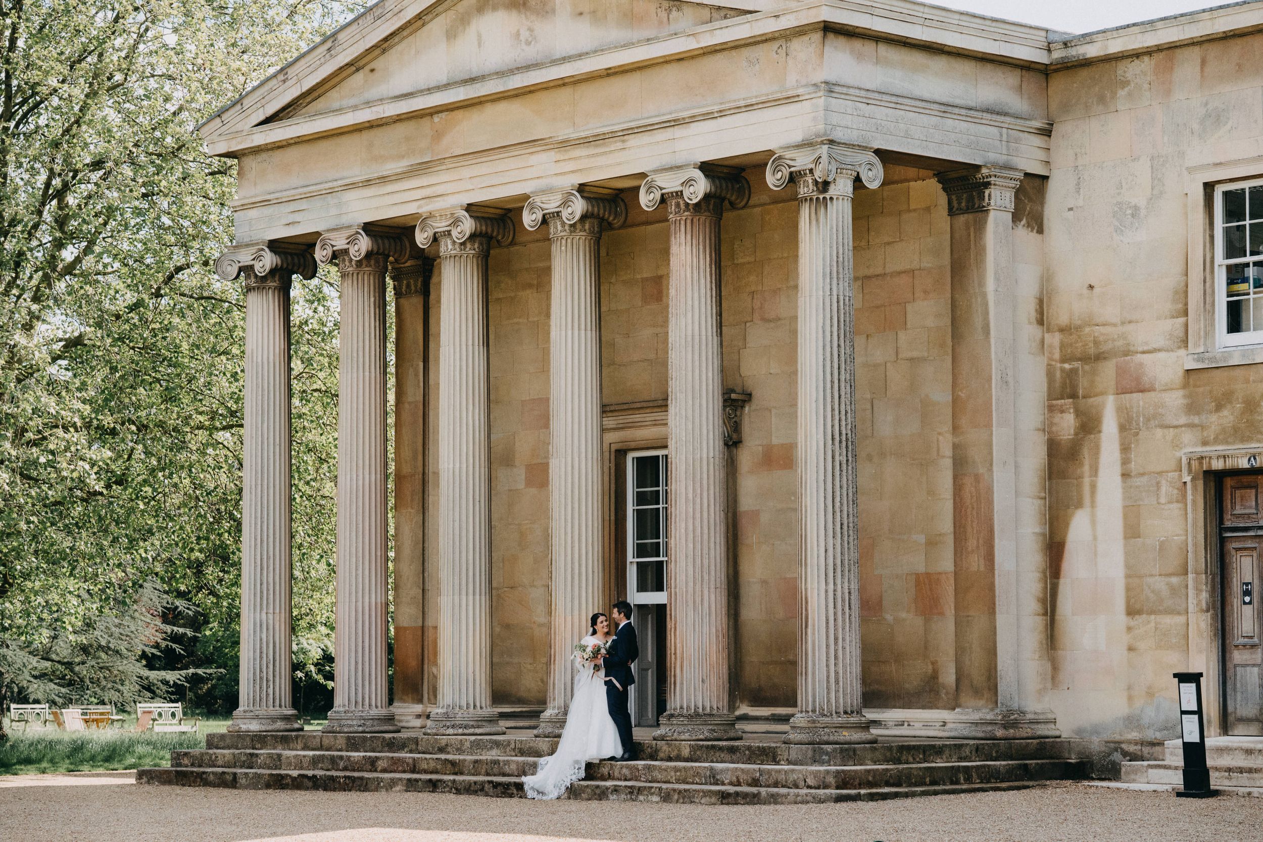Wedding couple standing by massive stone columns of a neoclassical building in Downing College Cambridge.