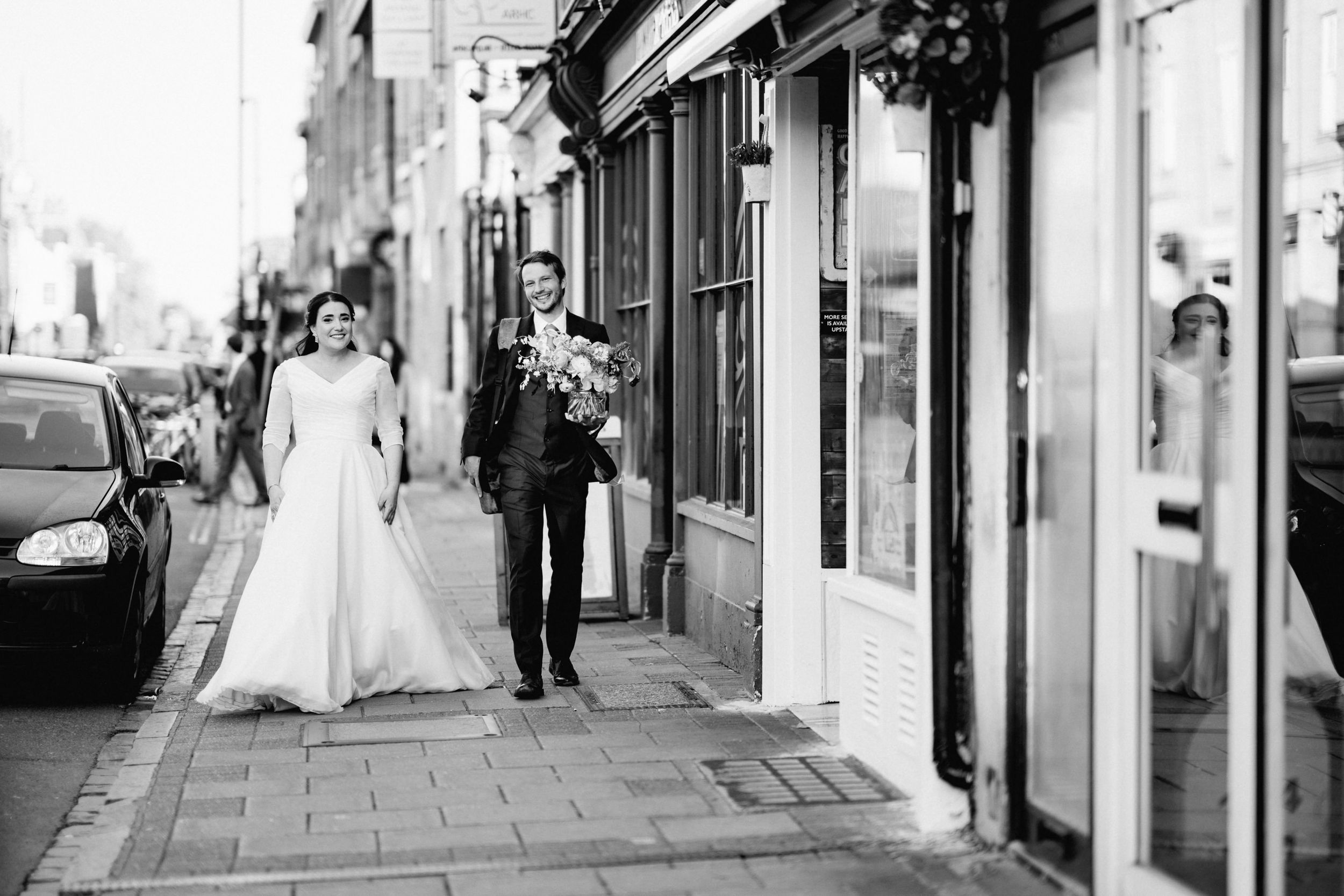 A black and white wedding photo of a couple walking along a charming city street of Cambridge with storefronts.