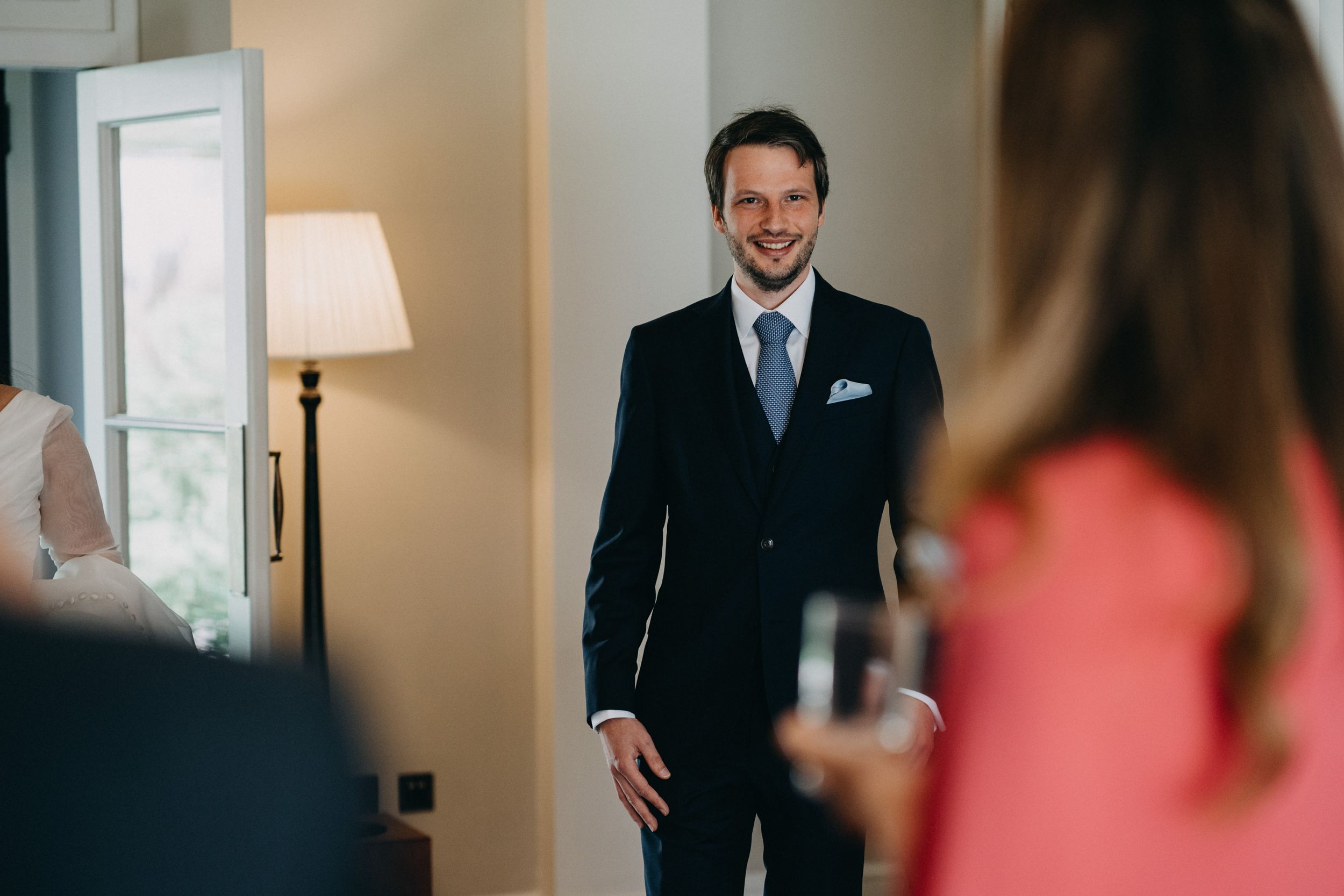 A groom in a dark suit and blue tie smiles while standing in an elegant room with warm lighting.