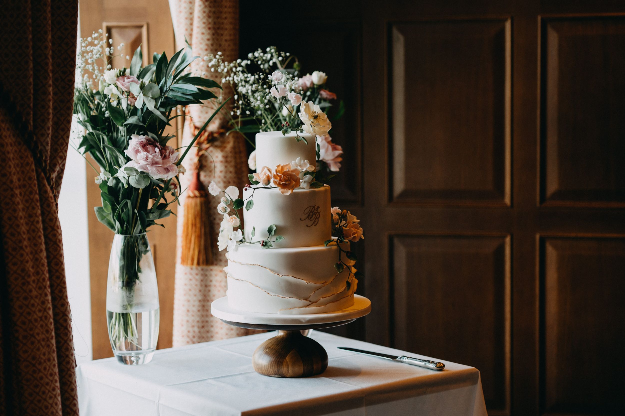 A tiered white wedding cake and floral arrangement displayed by a window with elegant tasseled curtains.