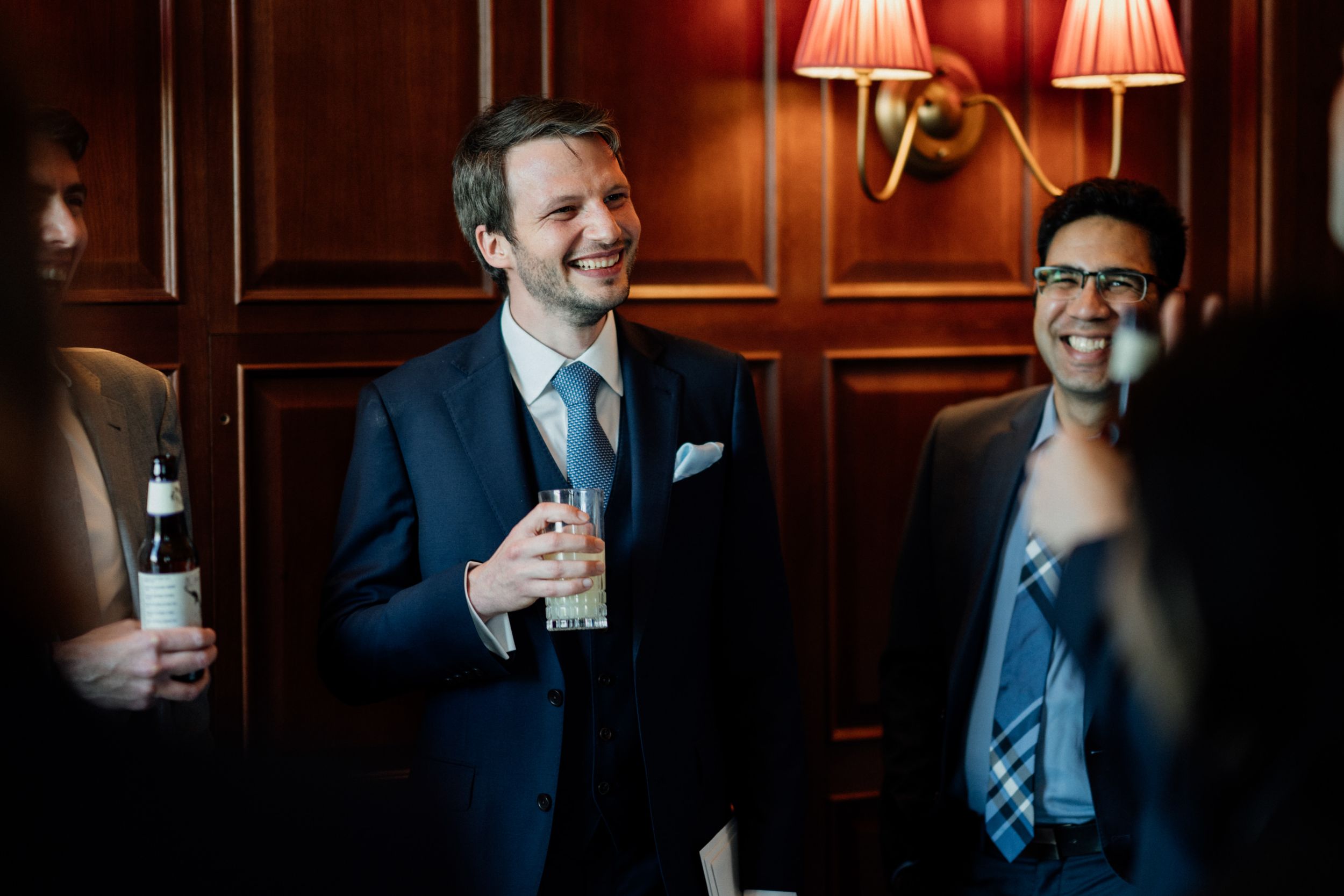Wedding guests enjoying drinks and conversation in an elegant wood-paneled room with red wall sconces.