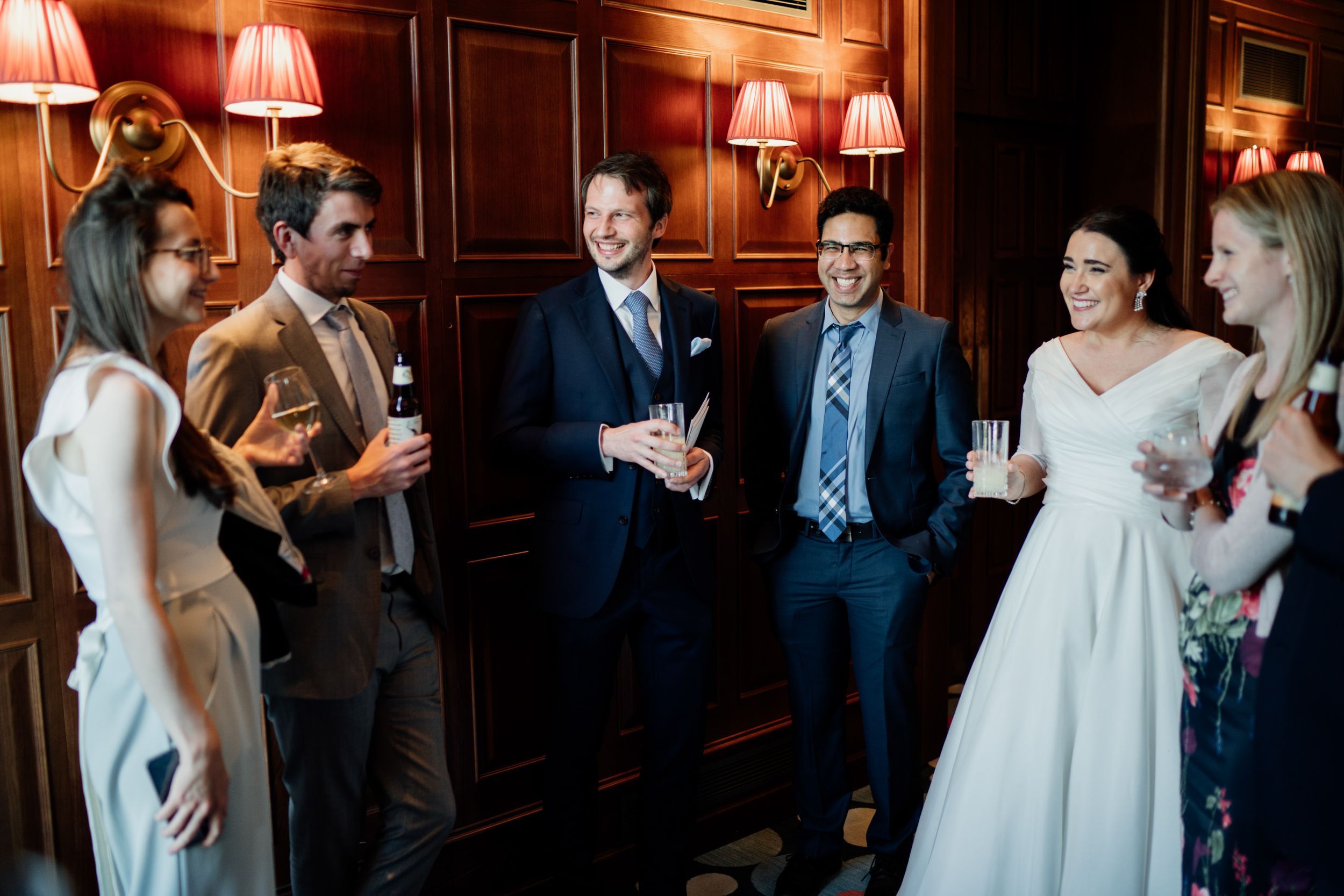 Wedding guests engage in conversation in an elegant wood-paneled room with warm lighting.