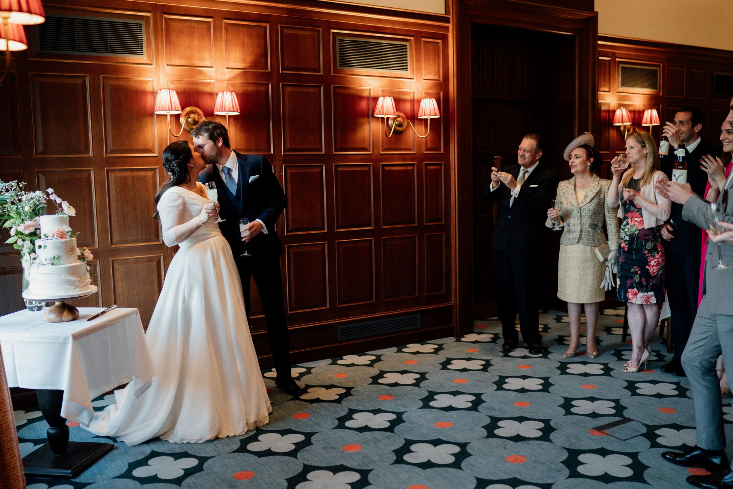 Newlyweds cut their wedding cake while guests watch and take photos in an ornate reception venue.