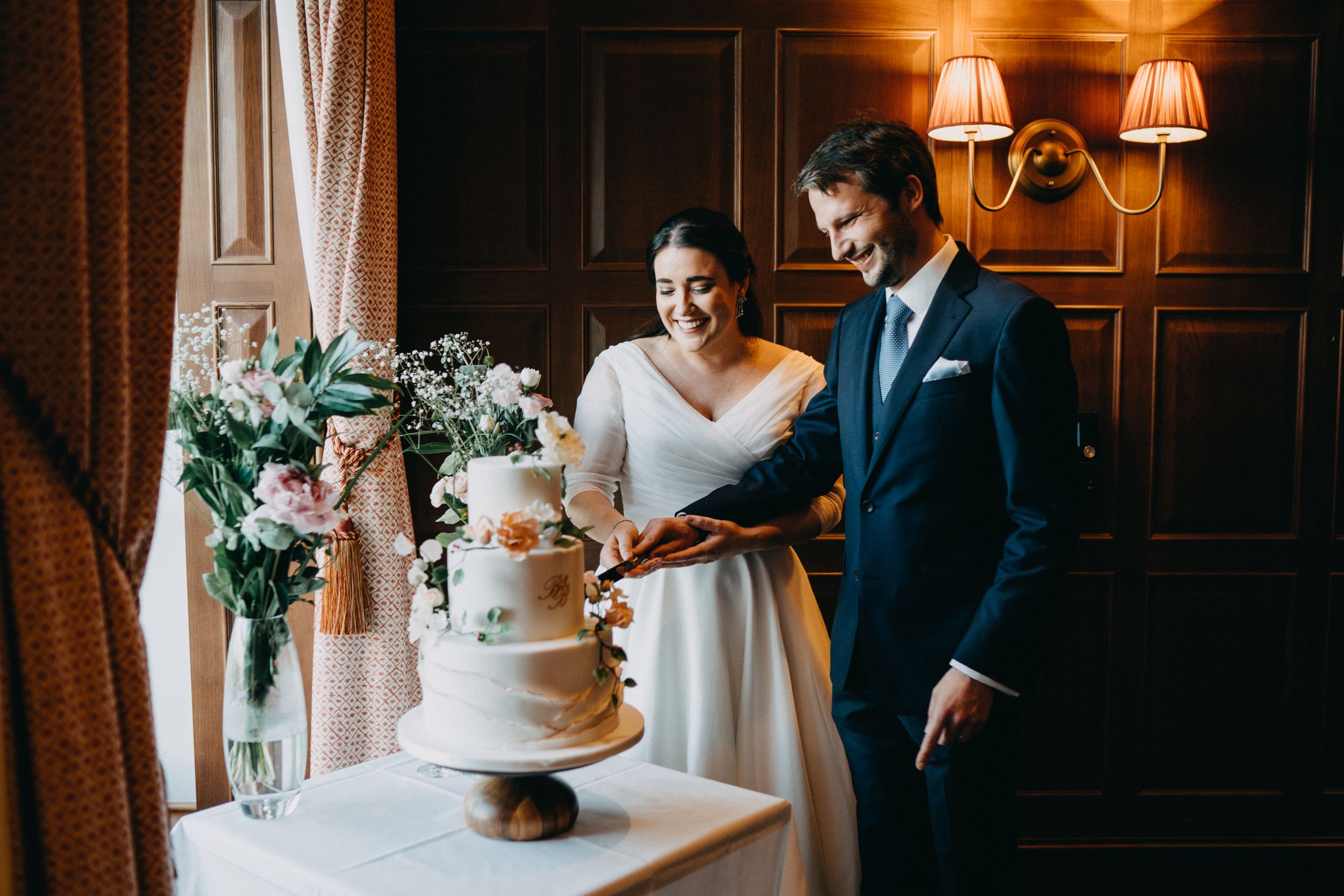 Couple stands next to their elegant three-tier wedding cake with floral decorations at University Arms Hotel, Cambridge.