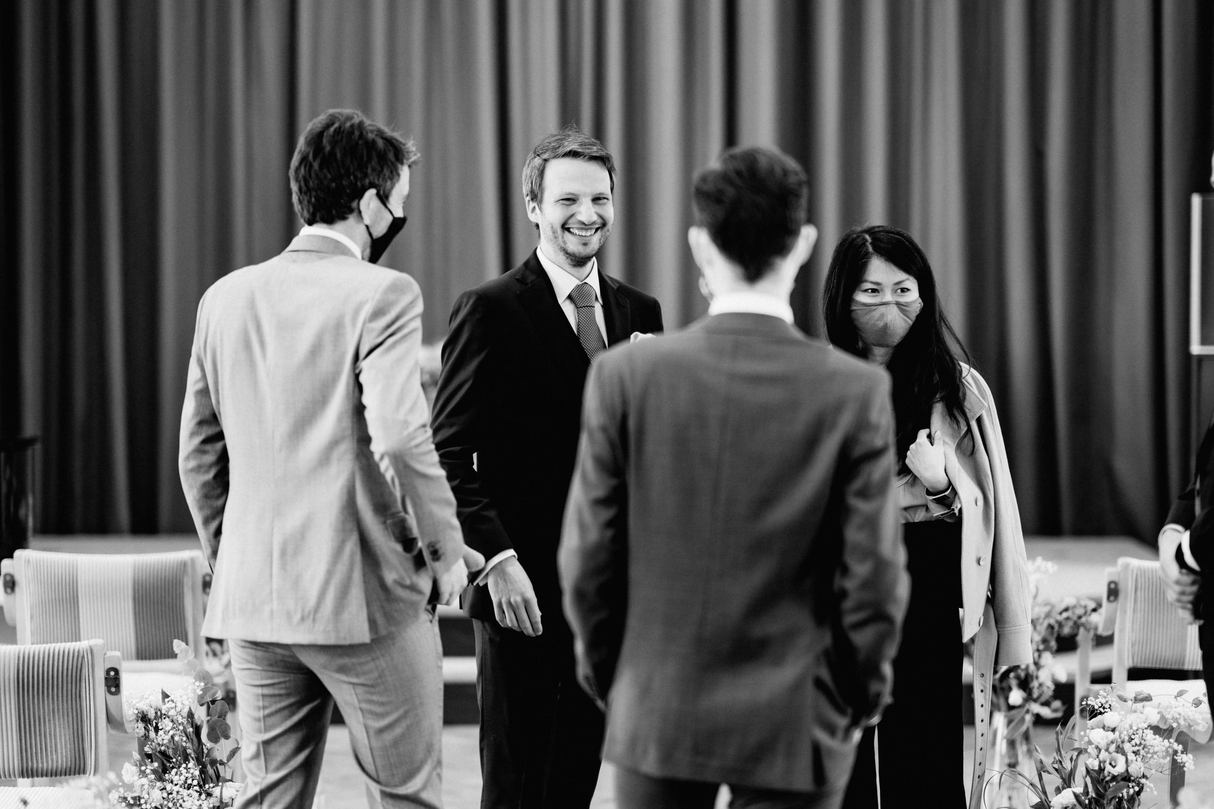 Wedding guests engage in conversation at an indoor reception with curtained backdrop.