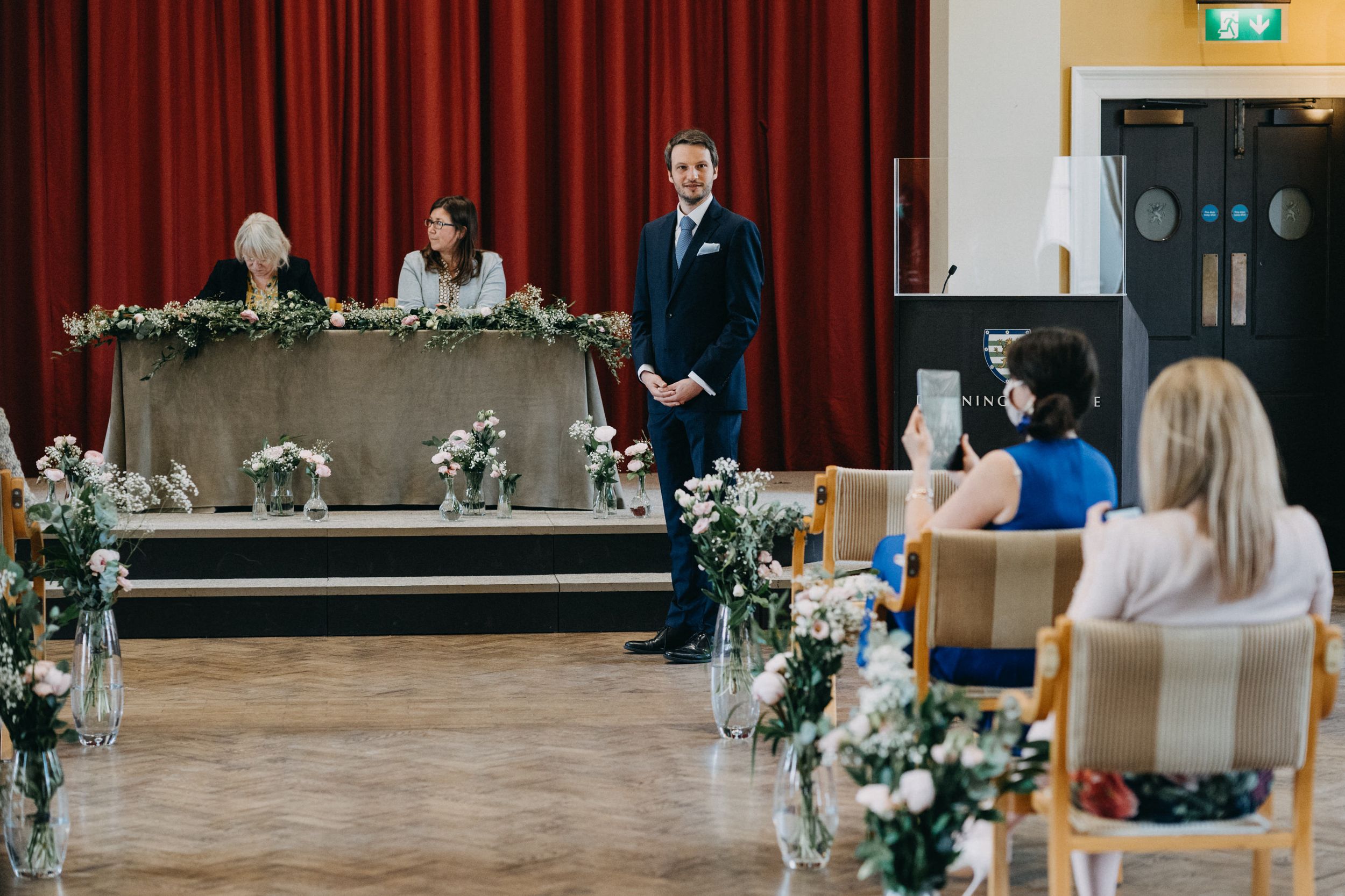 An elegant wedding ceremony setup with floral decorations and red curtains in a spacious hall of Downing College Cambridge.