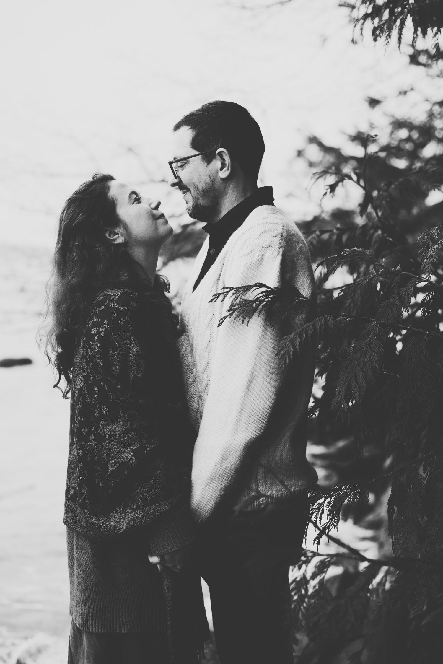 A couple stands closely together, smiling as they hold hands, with lush greenery framing the serene outdoor scene.