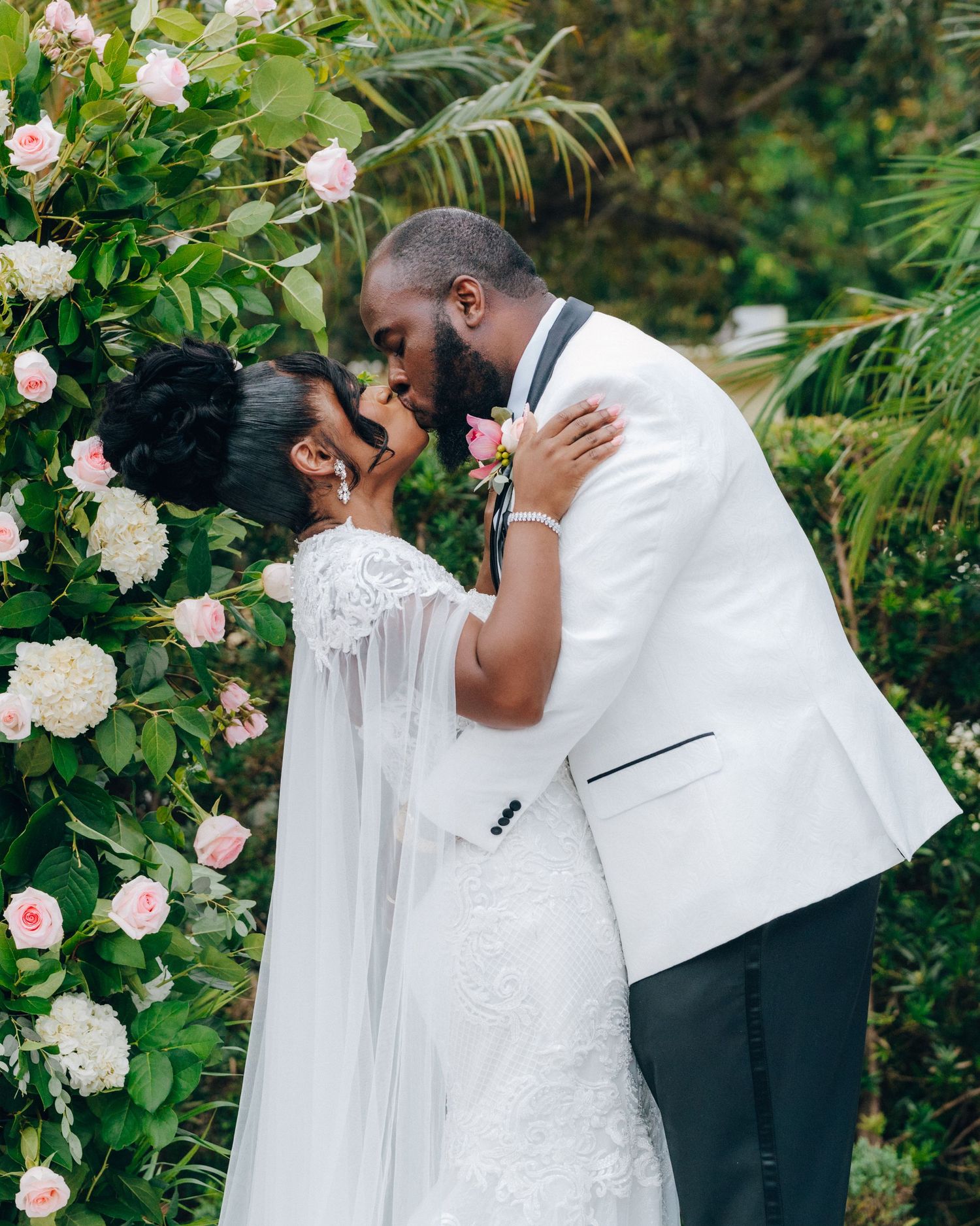 Romantic wedding moment captured among pink roses and tropical greenery.