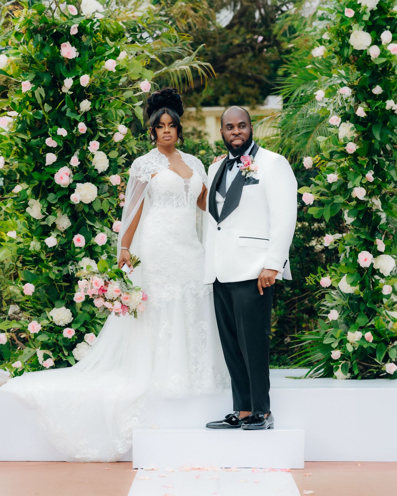 Elegant couple posing under lush floral arch with white roses and greenery.