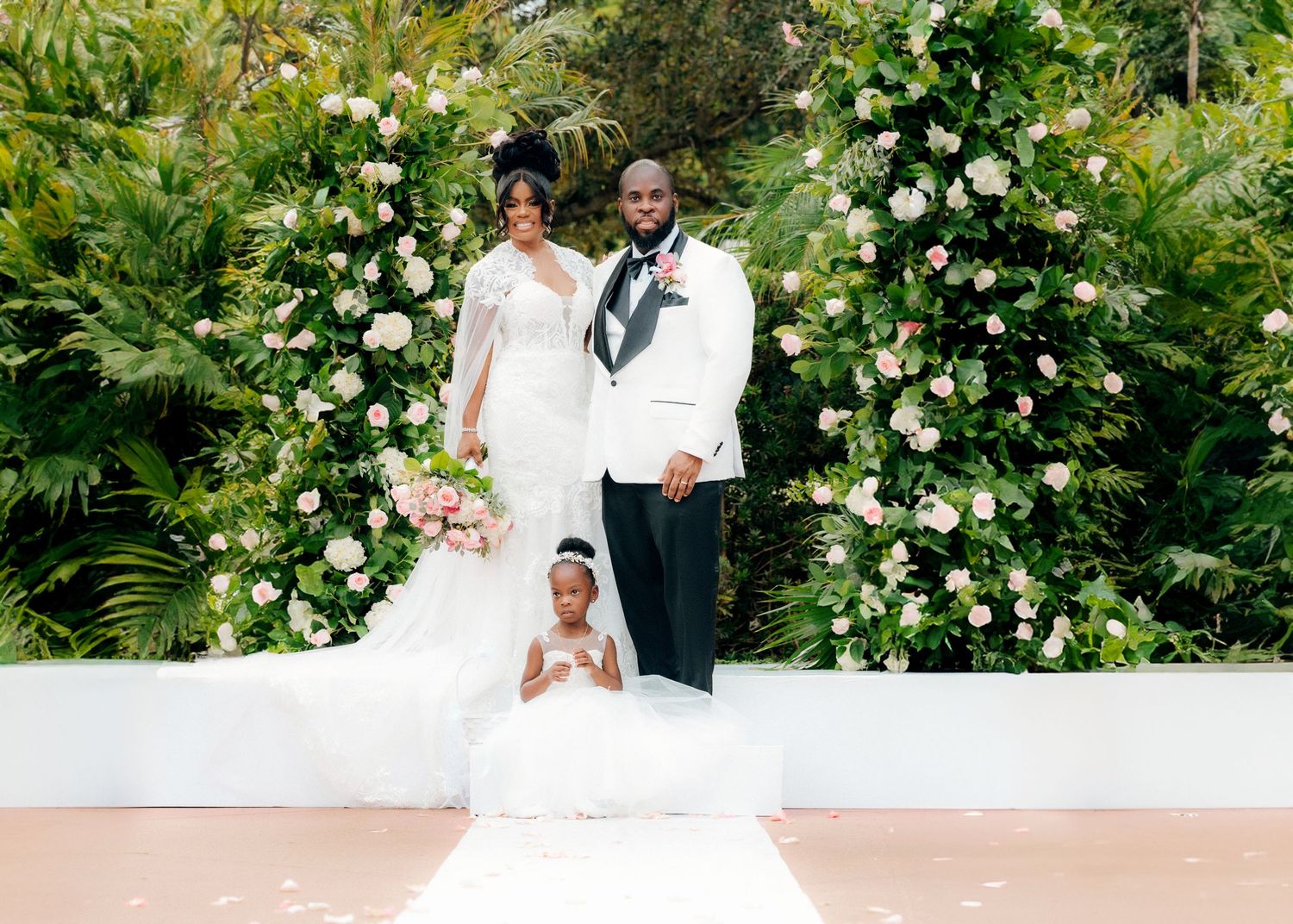 Wedding portrait under floral arch with white roses and tropical greenery.