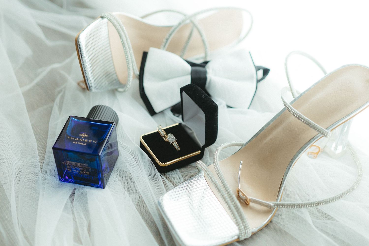 Wedding accessories including white bow tie, blue perfume bottle, ring box and bridal shoes on white tulle fabric.