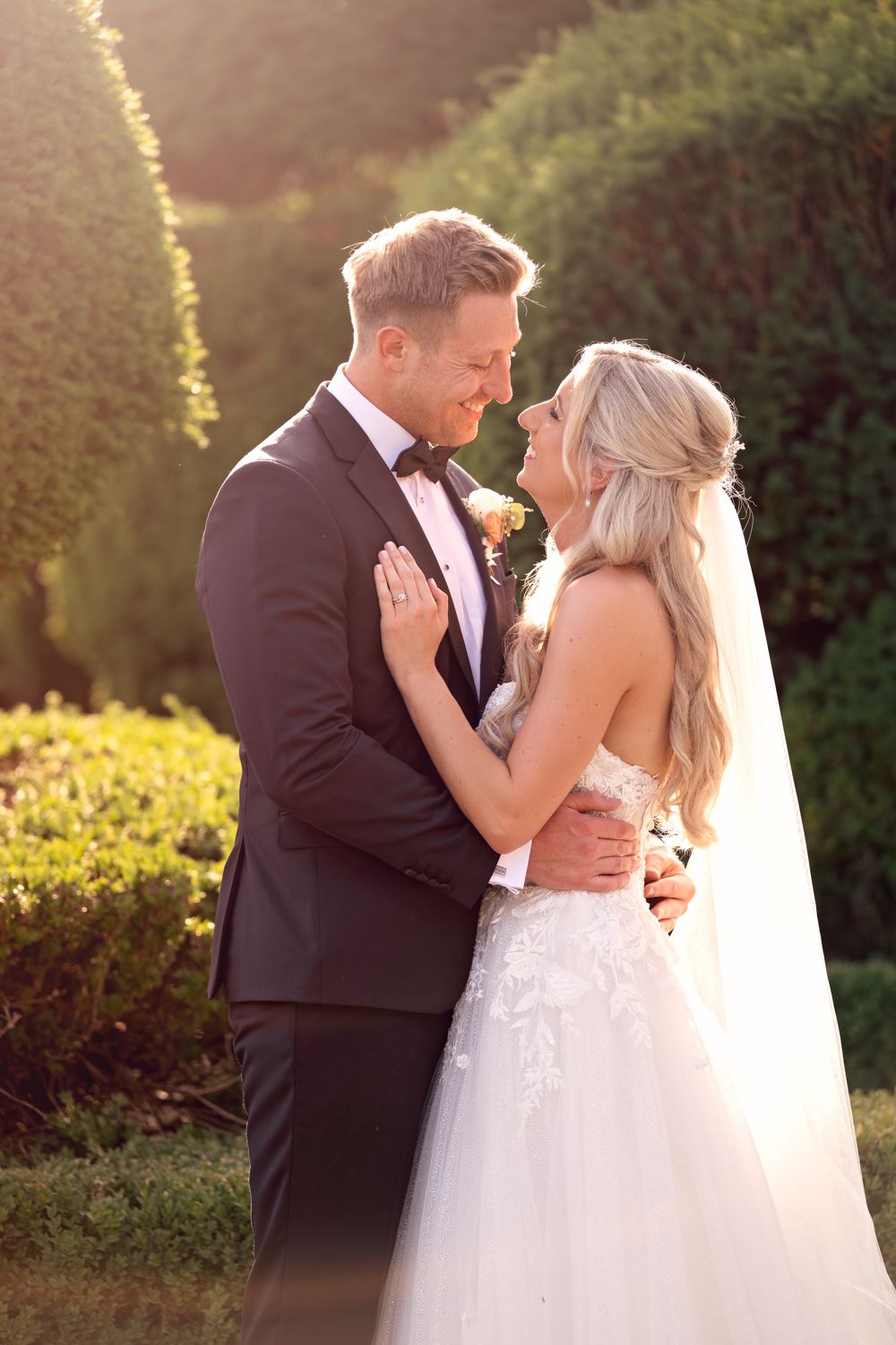 Newlyweds embrace in a romantic sunset garden setting with the bride wearing a white tulle gown and long veil.
