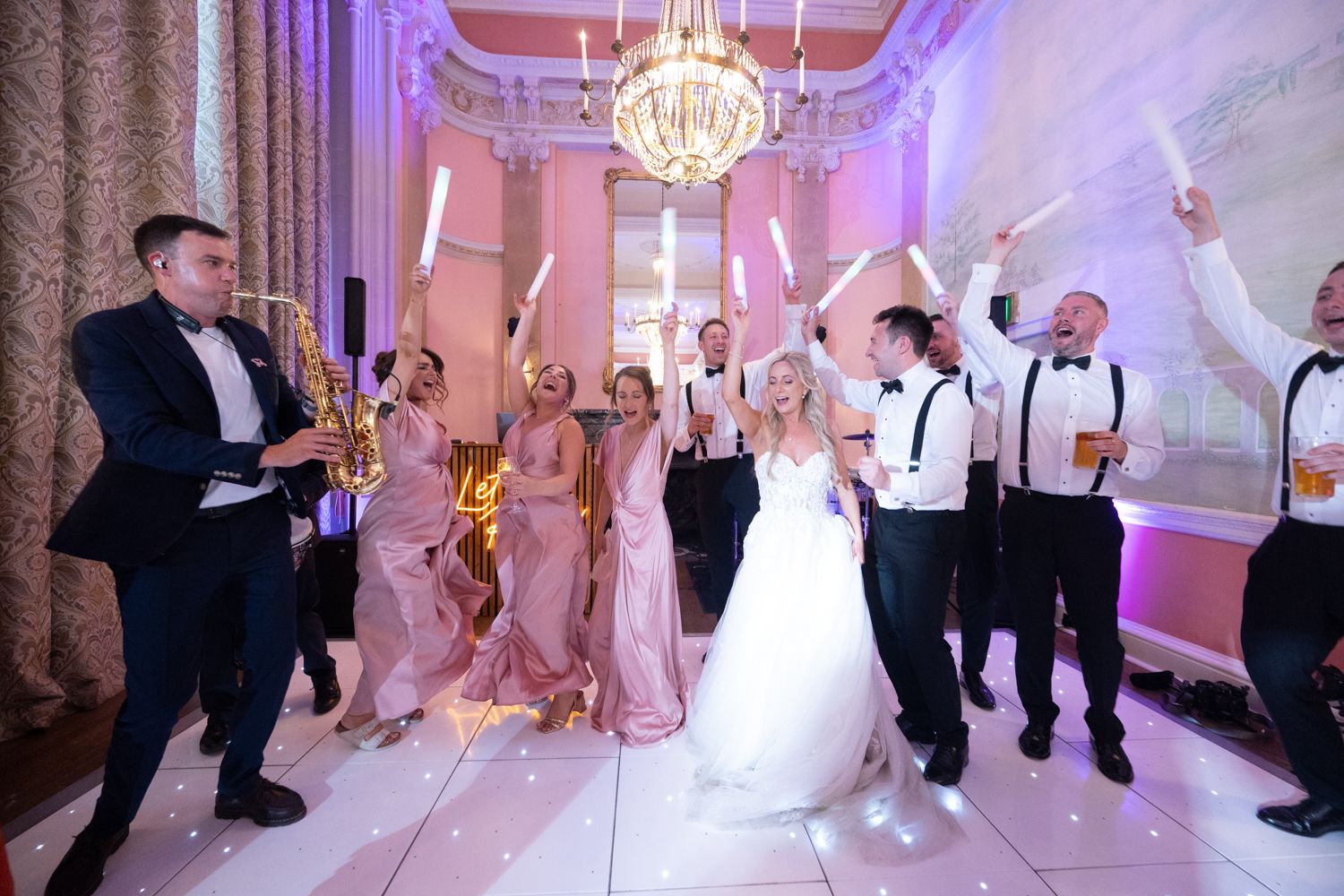 Wedding party dancing and celebrating on an illuminated dance floor in an elegant ballroom with purple lighting.