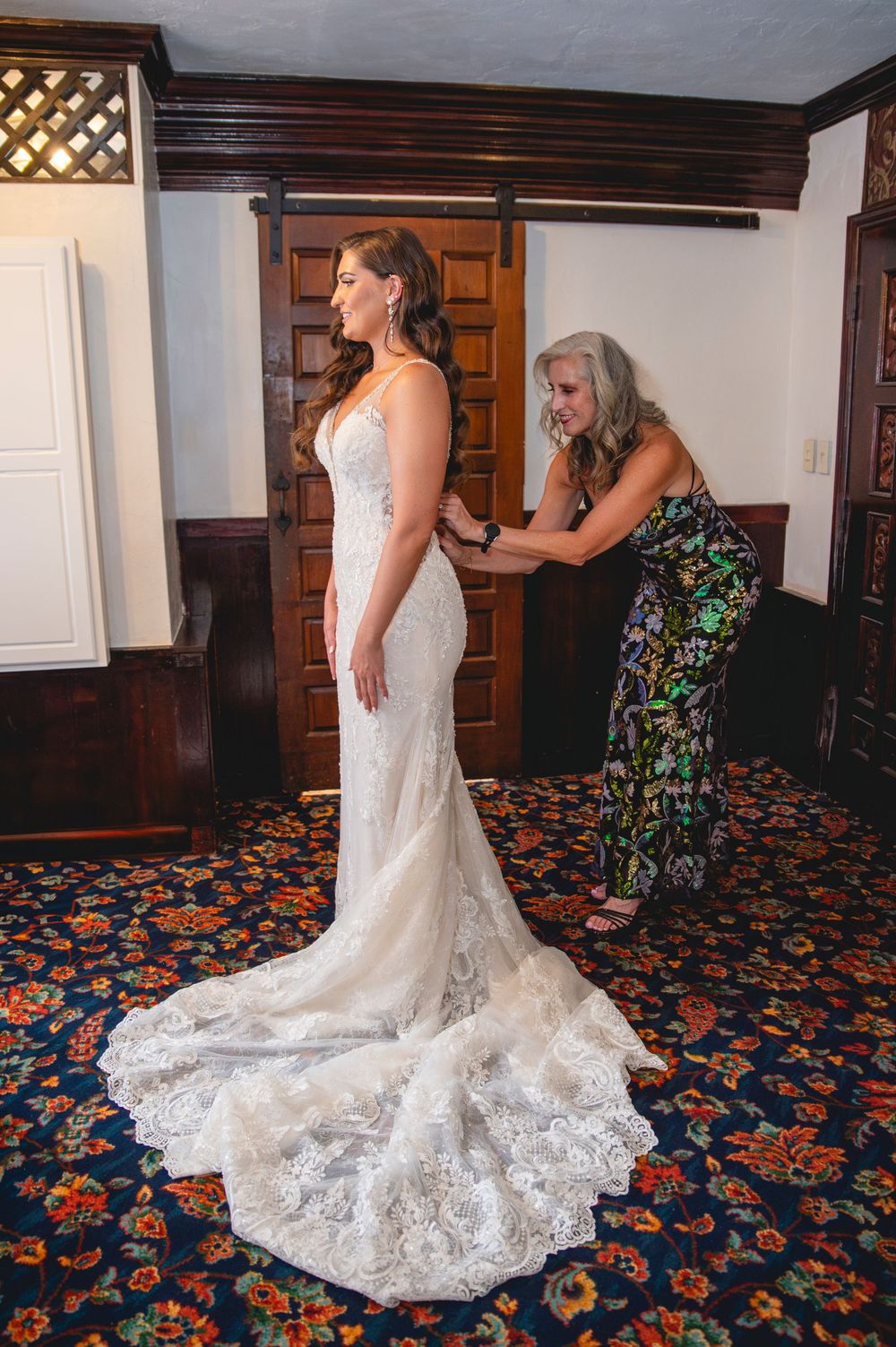 A bride in a white lace wedding dress with a flowing train stands on patterned carpet while being helped with final adjustments.