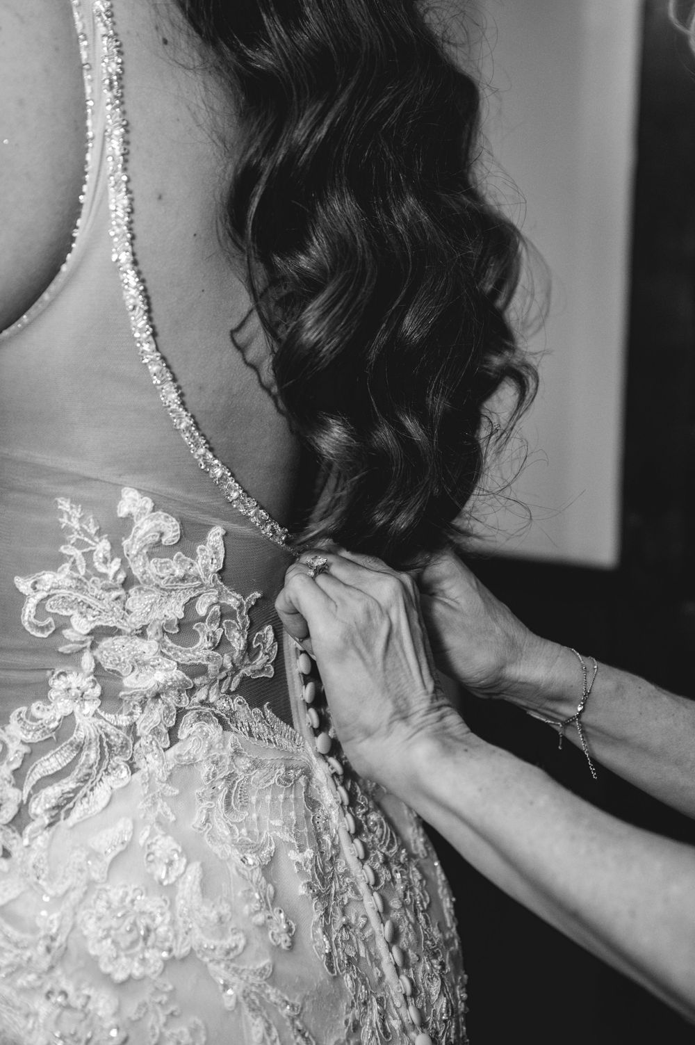 Hands adjust an intricate lace wedding dress detail with delicate beading and embroidery in a black and white photograph.