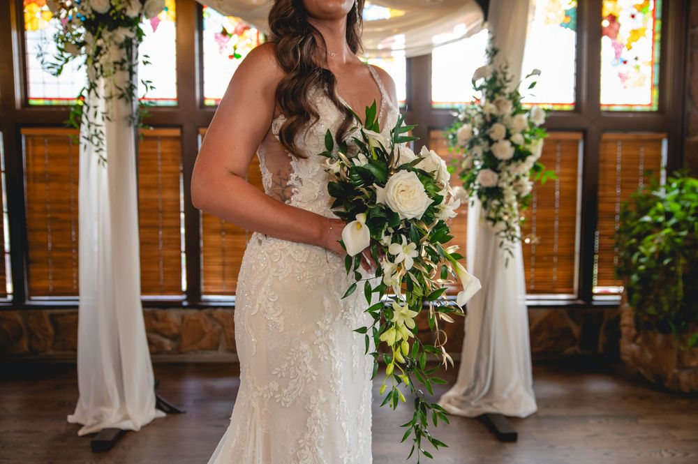 A wedding party in white dresses and formal attire stands in a rustic venue with floral decorations and natural lighting.