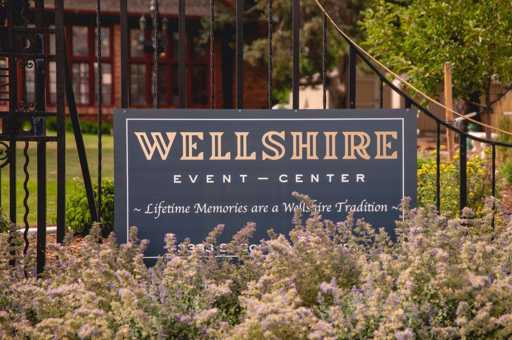 A rustic outdoor event center entrance with decorative cherry blossoms and a Wellshire sign among spring landscaping.