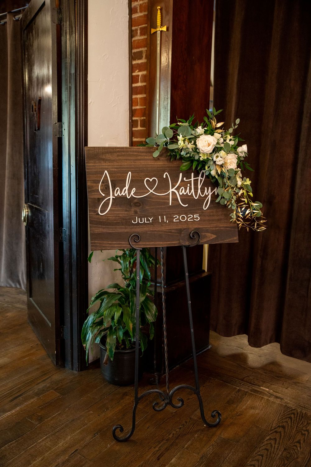 A wooden wedding welcome sign with white script lettering and floral decorations on an iron easel stand against a brick wall.