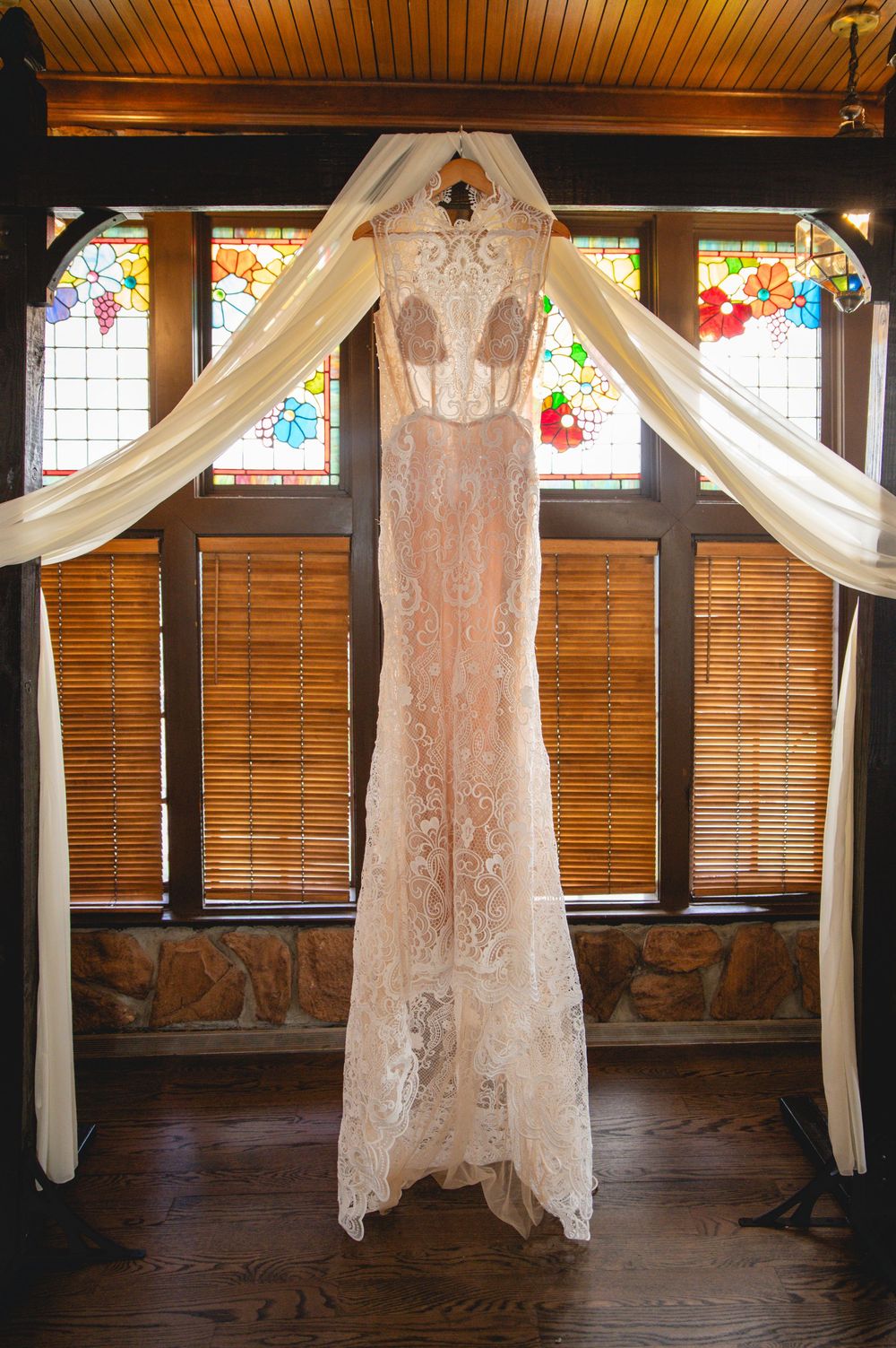 A delicate lace wedding dress hangs elegantly against white draping fabric and stained glass windows in a rustic wooden room.
