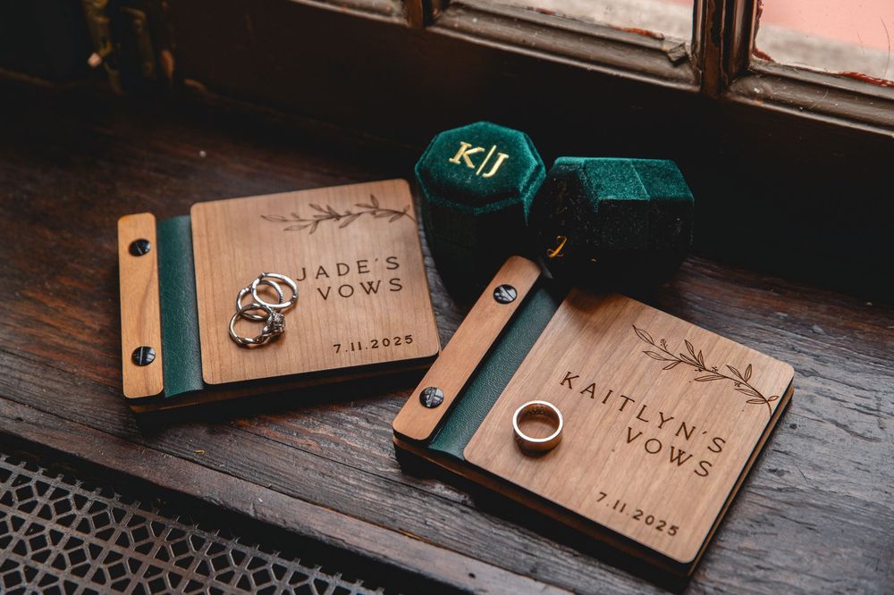 Two wooden wedding ring boxes with engraved names sit beside a green cube on a dark wooden surface.