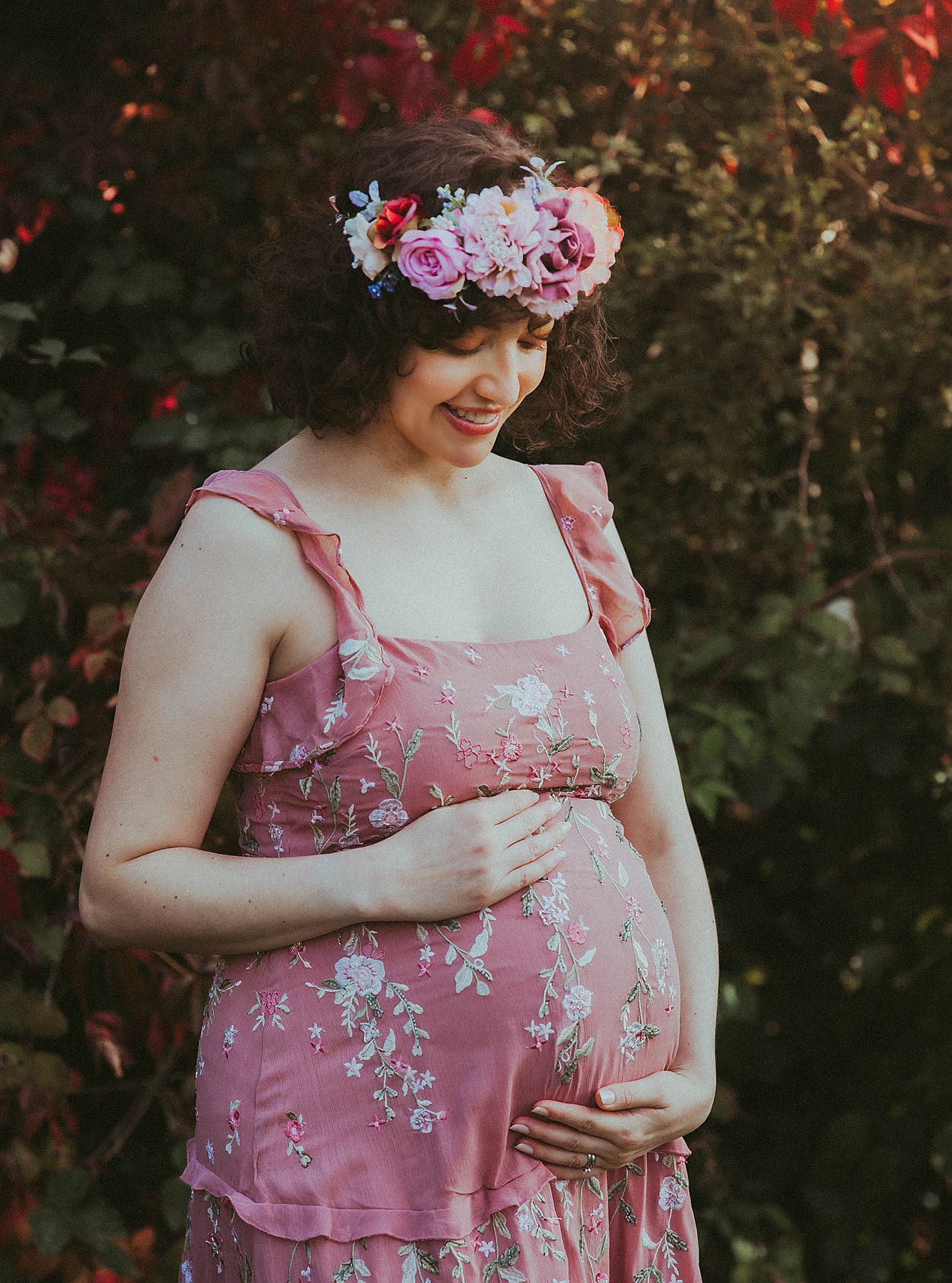 A pregnant woman in a pink floral dress and flower crown stands in front of red foliage during a maternity photo shoot.