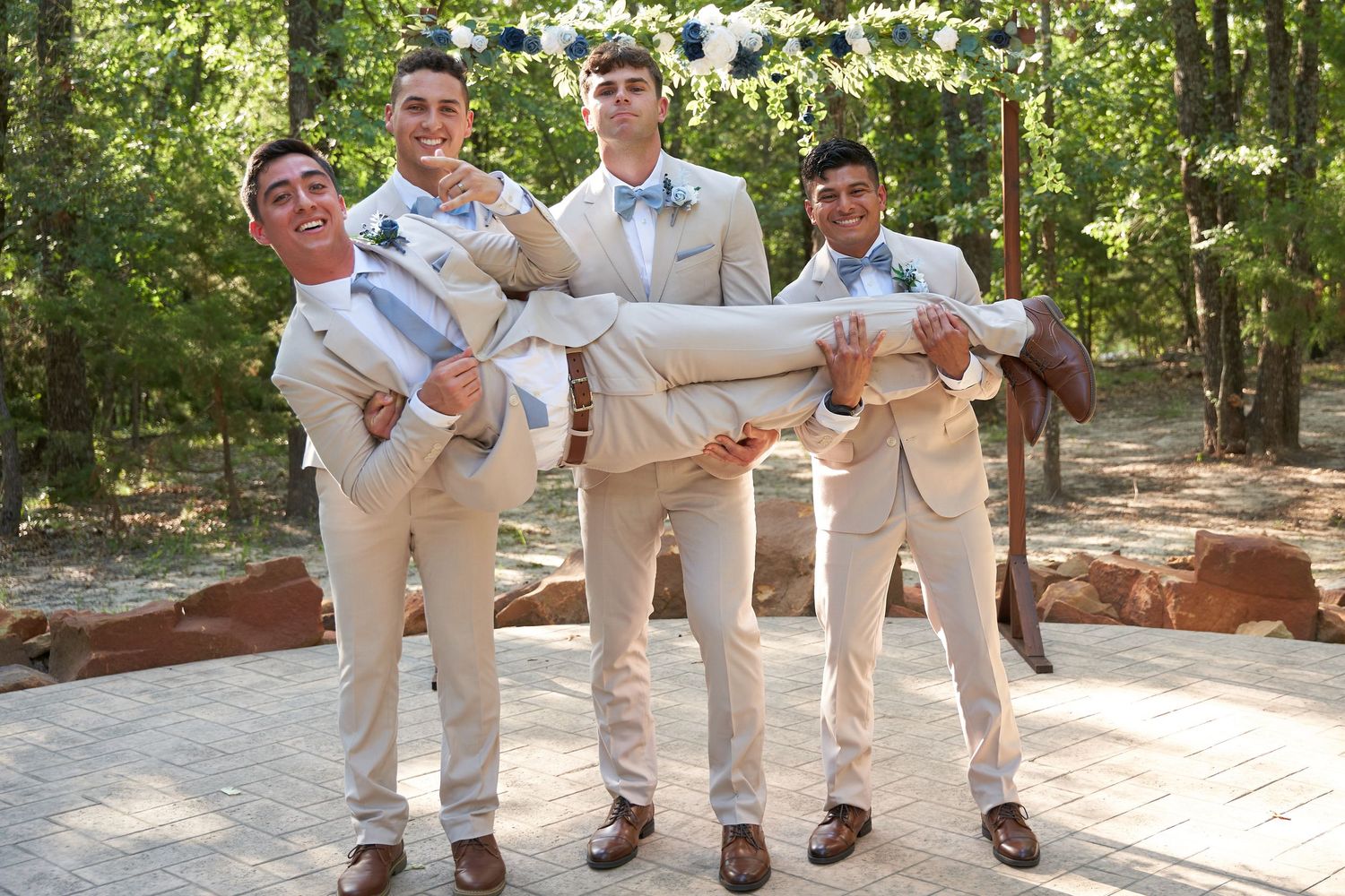 A group in light colored suits pose playfully in a wooded setting with sunlight filtering through the trees.