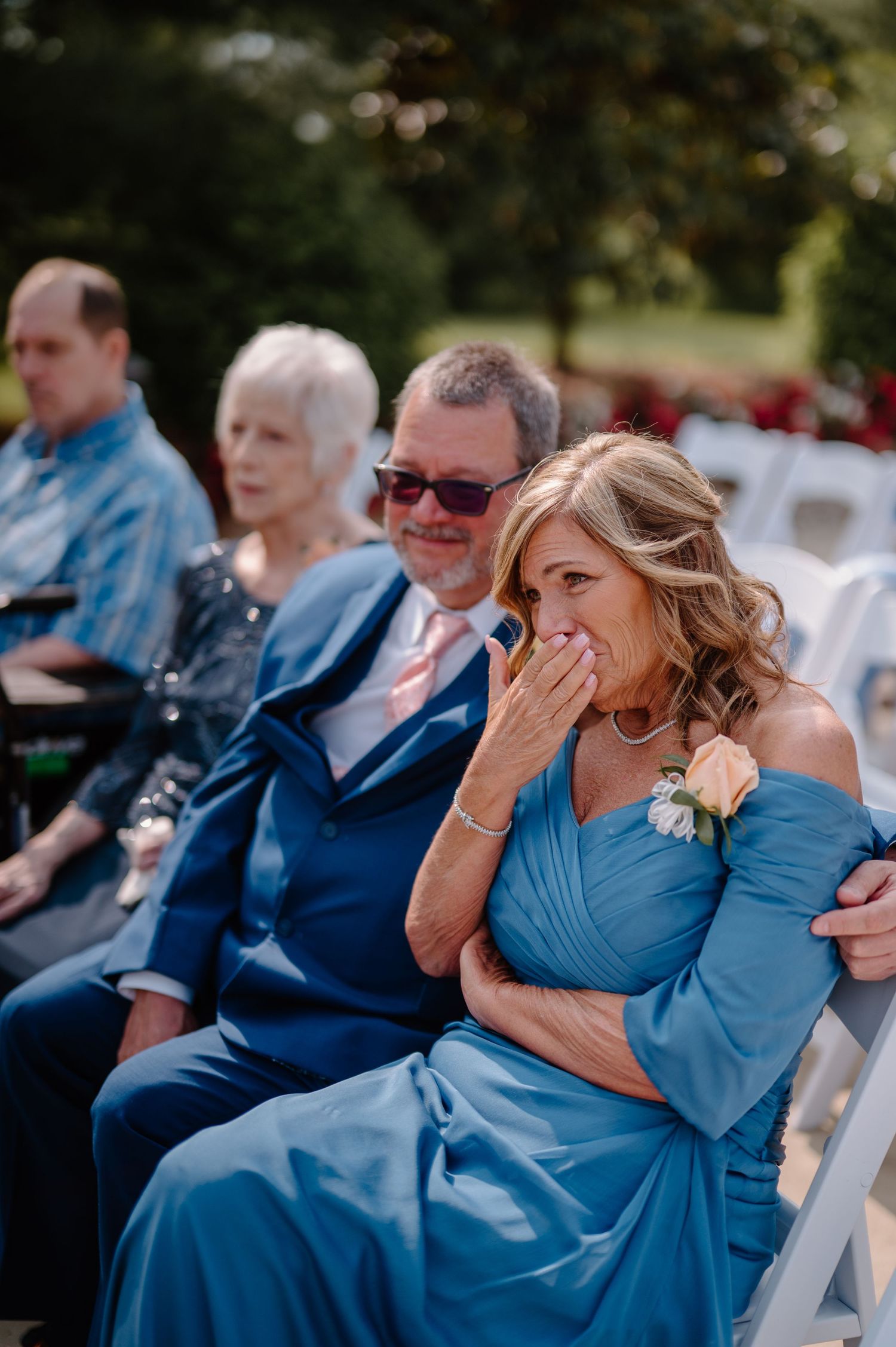 Four connected photos show emotional wedding guests in blue attire reacting during an outdoor ceremony.