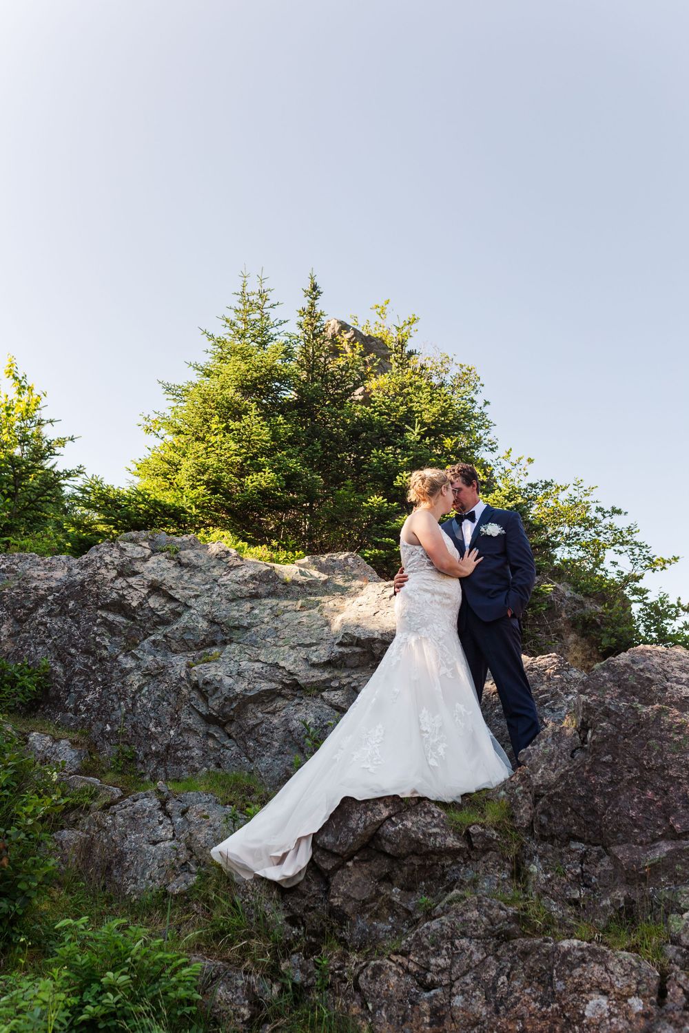 A romantic wedding couple embraces while standing on a rocky outcrop with evergreen trees in the background.