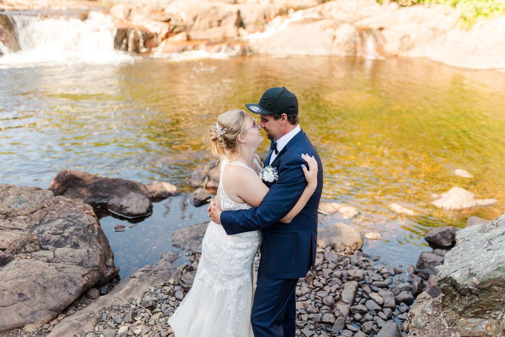 A couple in wedding attire embraces near a scenic waterfall with golden sunlit water and rocky terrain.