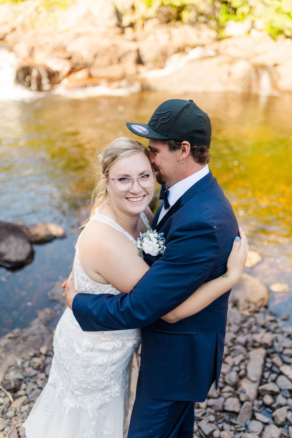 A newlywed couple shares romantic moments by a riverside with rocks and golden sunlight filtering through the trees.