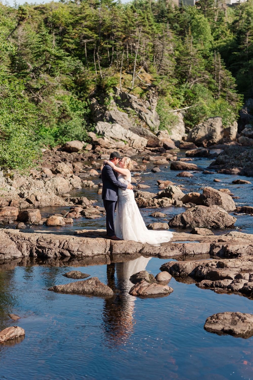 Two people embrace on rocky cliffs overlooking a flowing river surrounded by lush forest scenery on a sunny day.