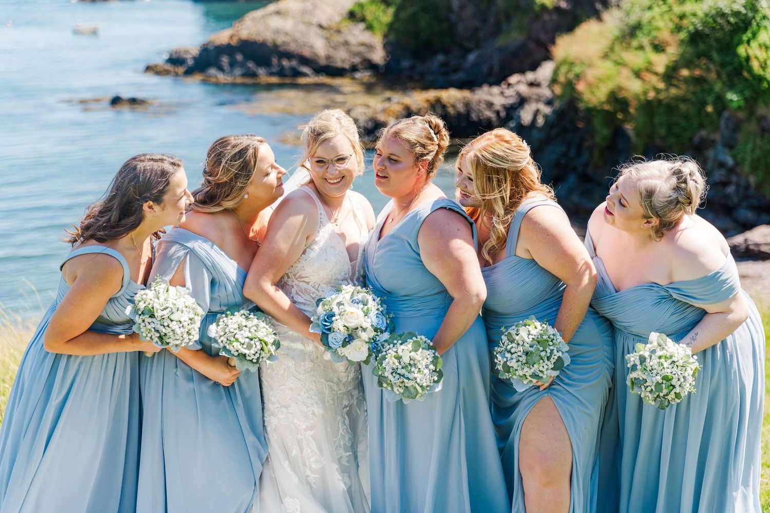 Wedding party in light blue bridesmaid dresses poses together by ocean coastline on sunny day.