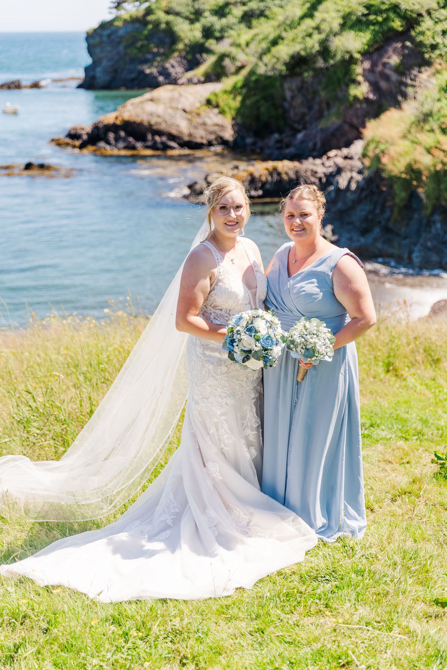 Wedding party stands on a grassy cliff overlooking a stunning coastal cove with blue water and rocky shoreline.