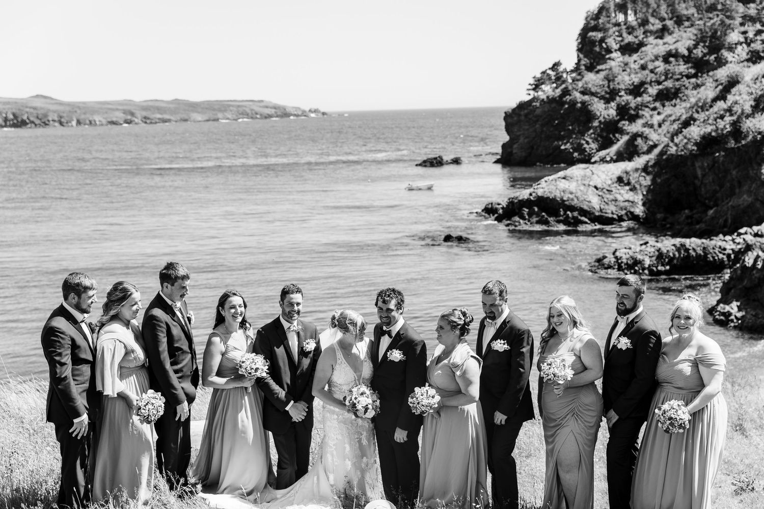 Wedding party stands in casual poses along a shoreline with ocean and cliffs visible in the black and white photo.