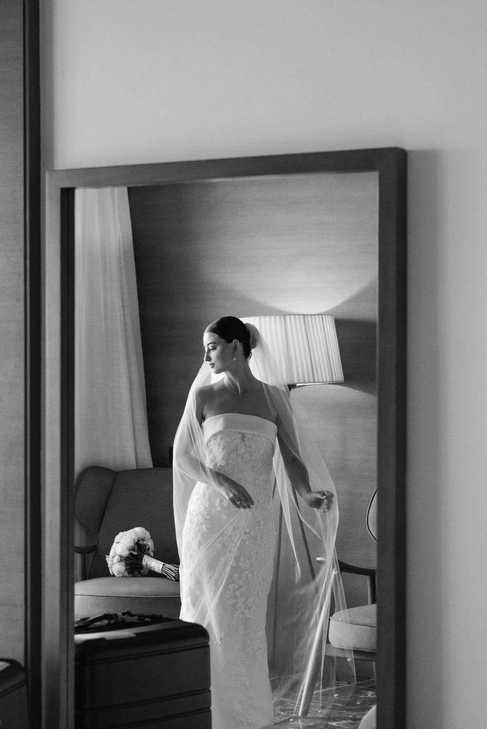 A black and white photo of a bride in a lace wedding dress looking in a mirror before her ceremony.
