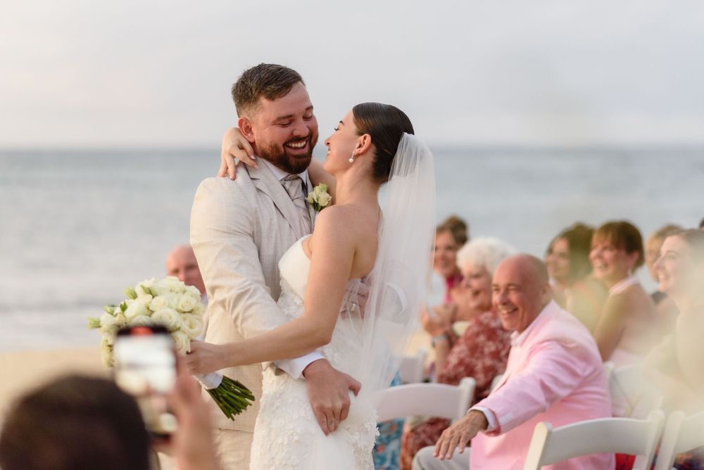 A couple shares an intimate moment during their beach wedding ceremony with guests watching from their seats by the ocean.