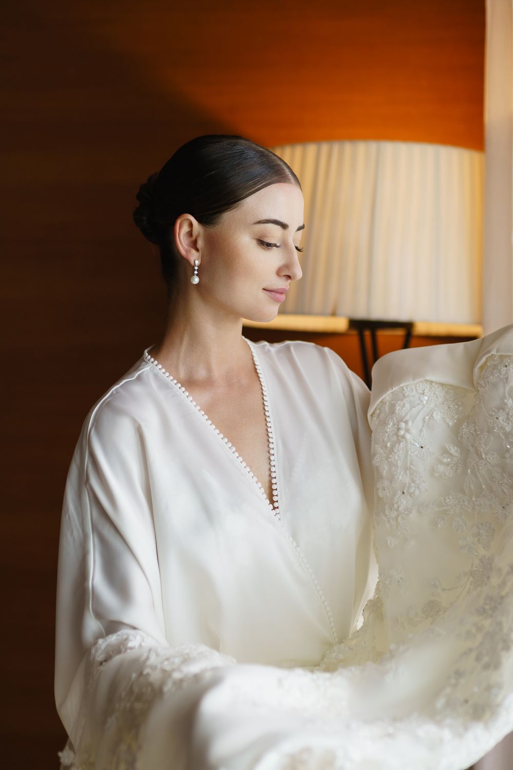 A bride wearing a white satin robe with decorative trim gazes down serenely during wedding preparations.