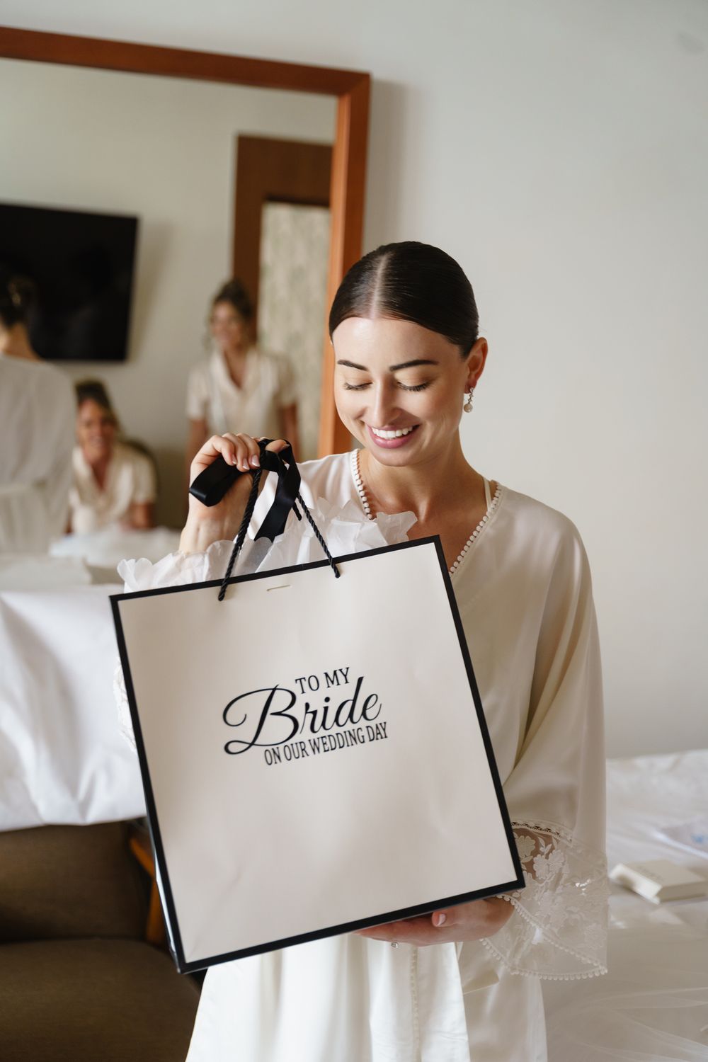 Smiling person in white robe holds a black and white wedding gift bag that says 'To my Bride' during preparations.