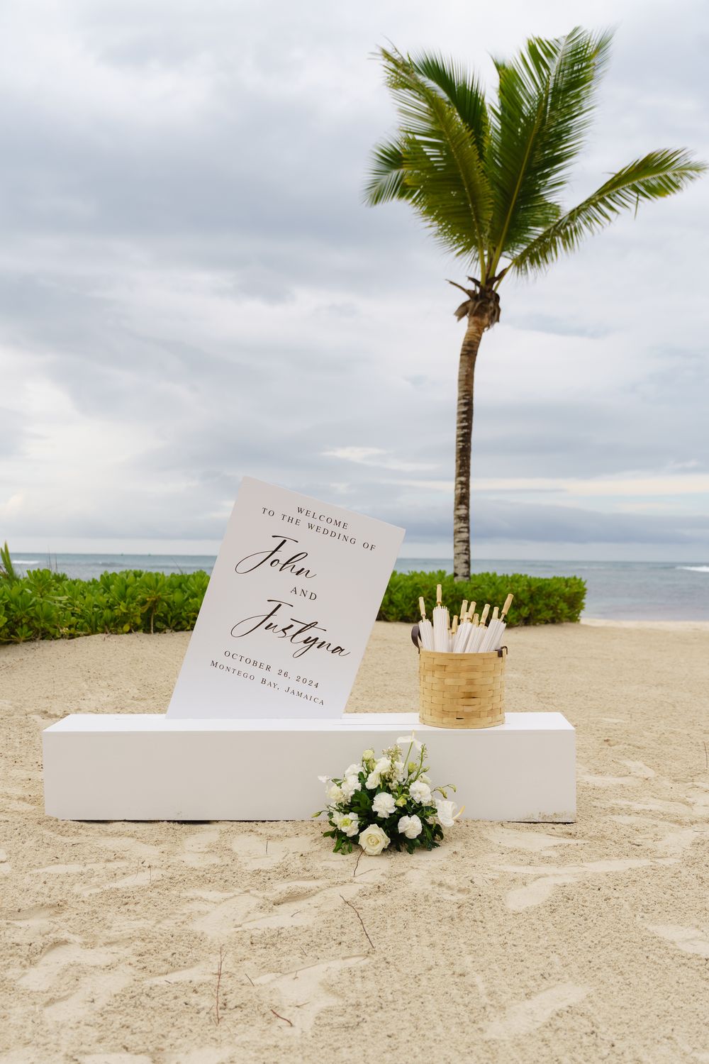 Wedding invitation display on a tropical beach with white floral arrangements and palm trees against a cloudy sky.
