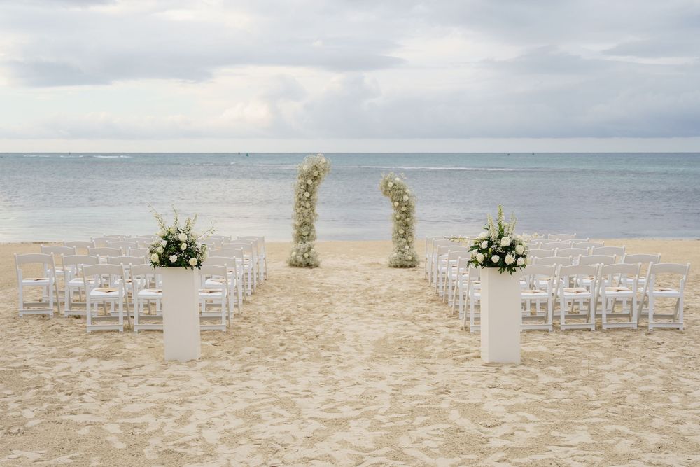A romantic beach wedding setup with white chairs and floral archway on the sandy shore overlooking the ocean.