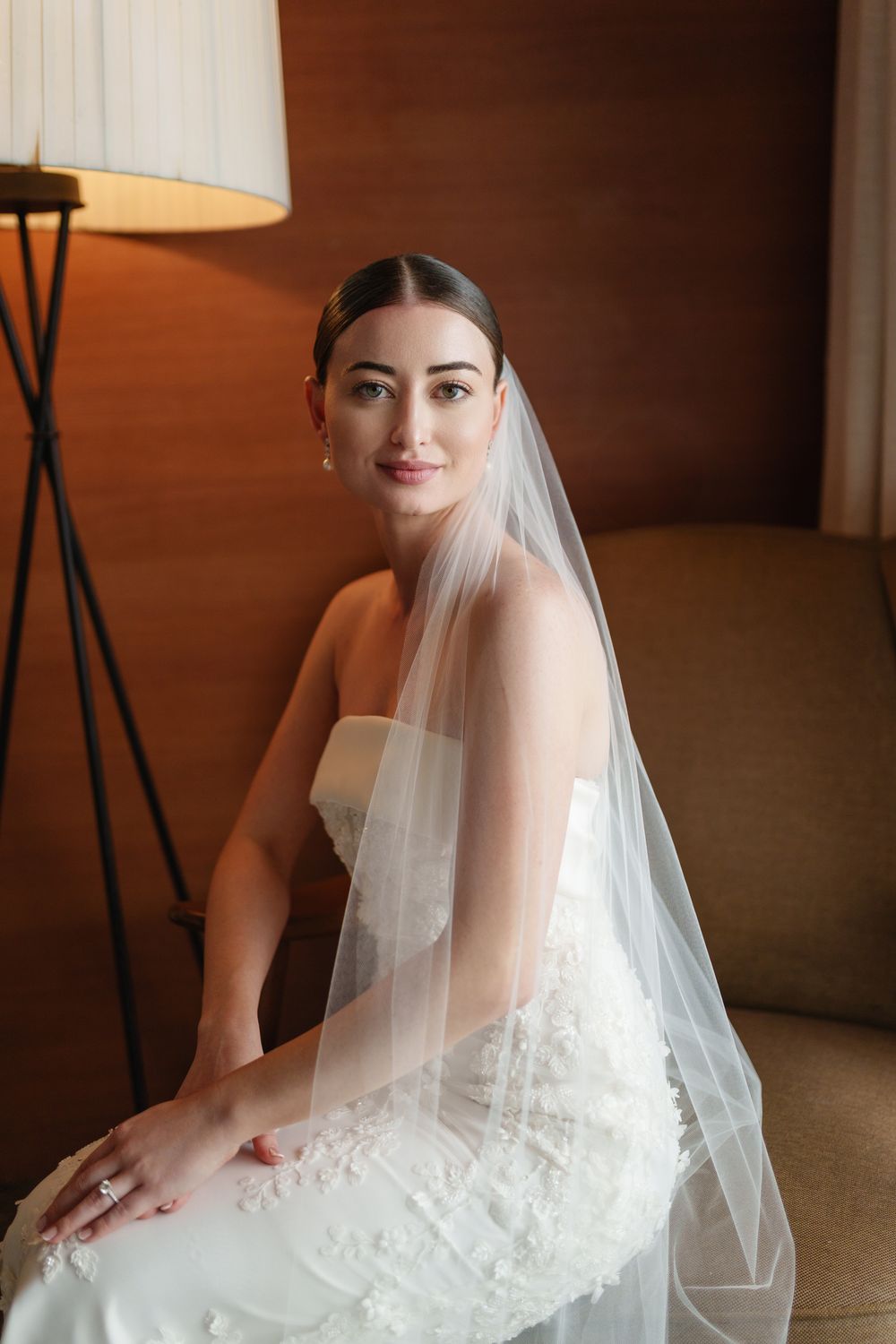 A bride in a strapless white wedding gown and veil sits elegantly in a room with warm lighting and a modern lamp.