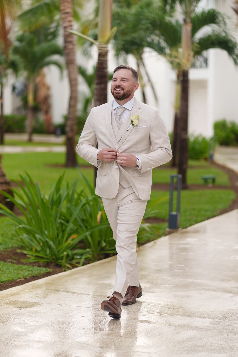 A groom in a beige suit and tie walks down a tropical palm-lined pathway.