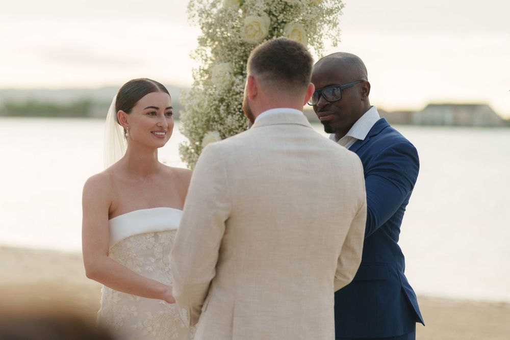 A beachside wedding ceremony takes place with a couple and officiant standing together at sunset.