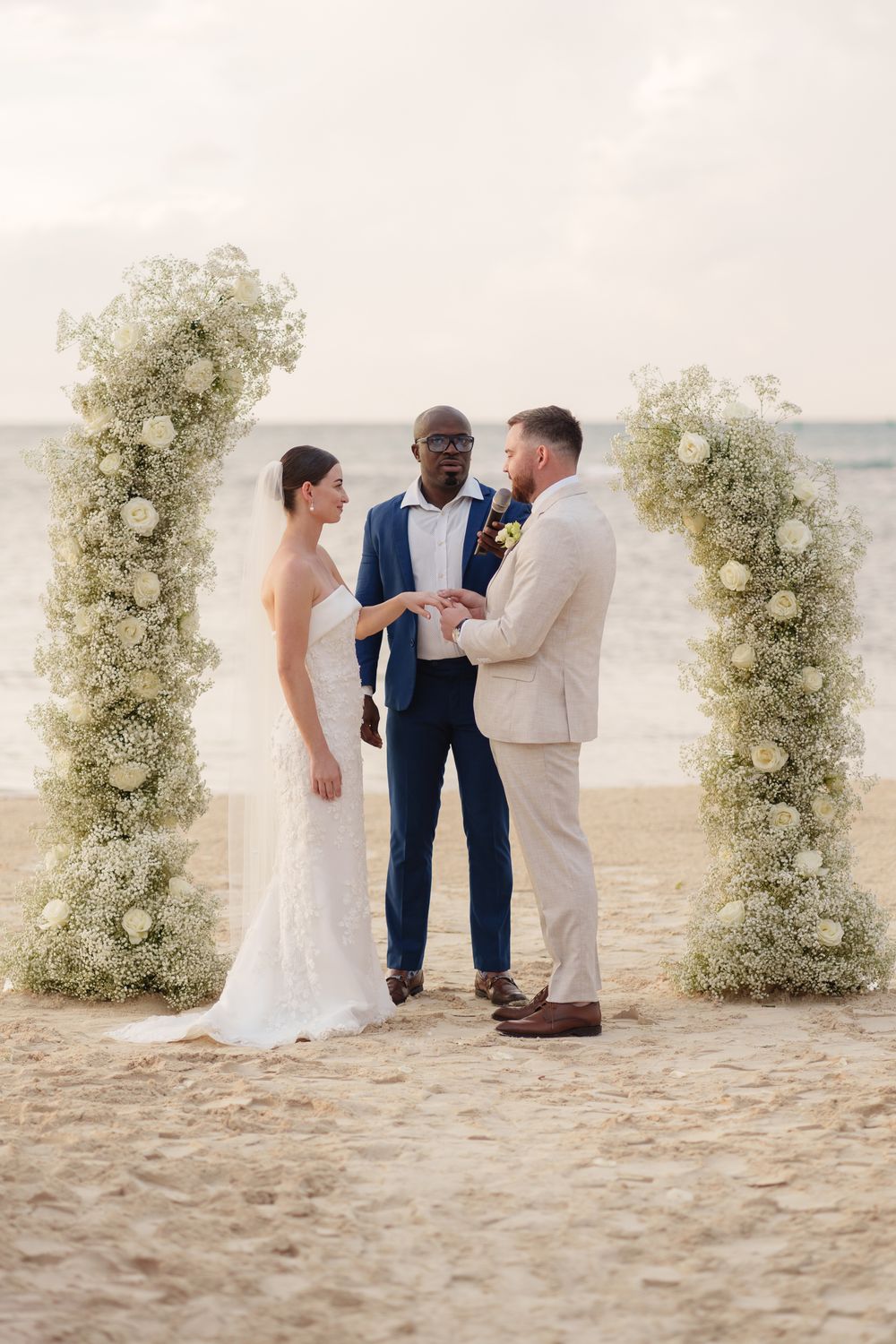 A beach wedding ceremony under floral arches with the couple and officiant on sand beside the ocean.