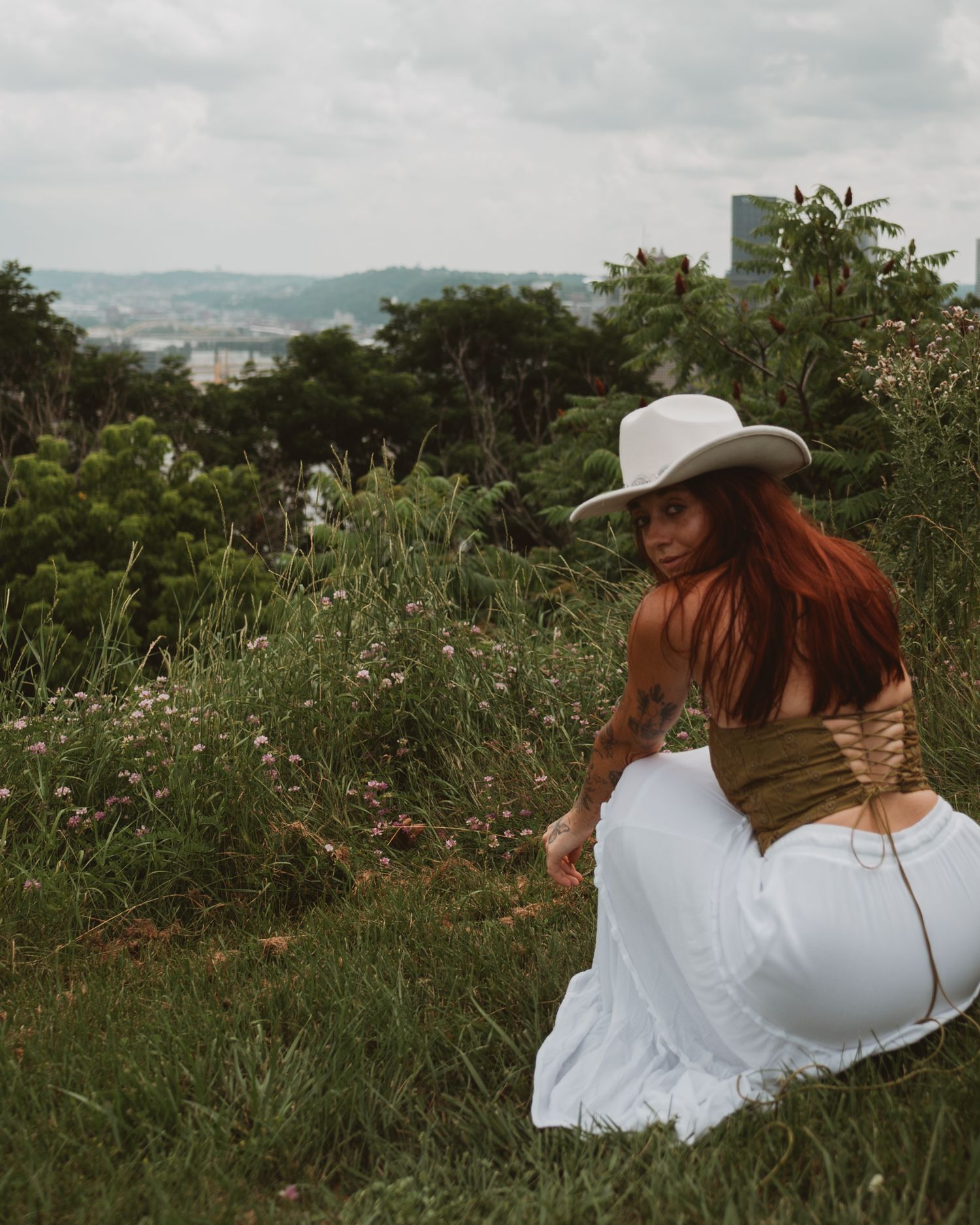 Figure in white dress and cowboy hat sits in grassy hillside meadow overlooking distant city view.