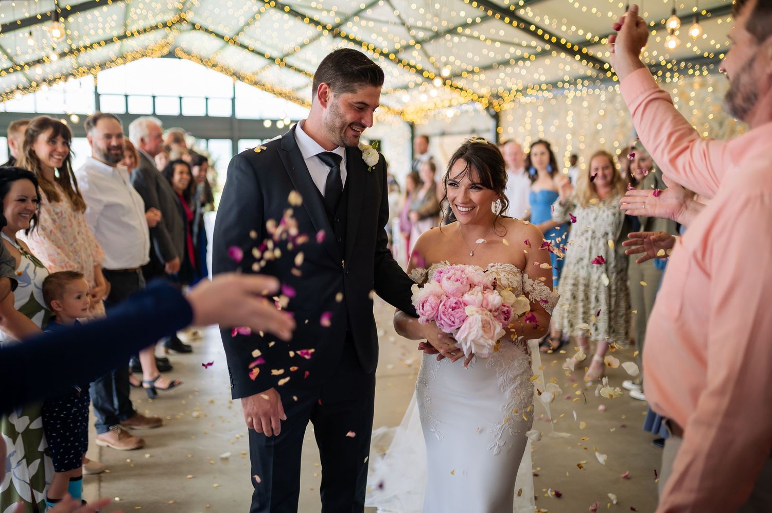 Wedding guests smiling and celebrating with confetti while walking under a tent decorated with twinkle lights.