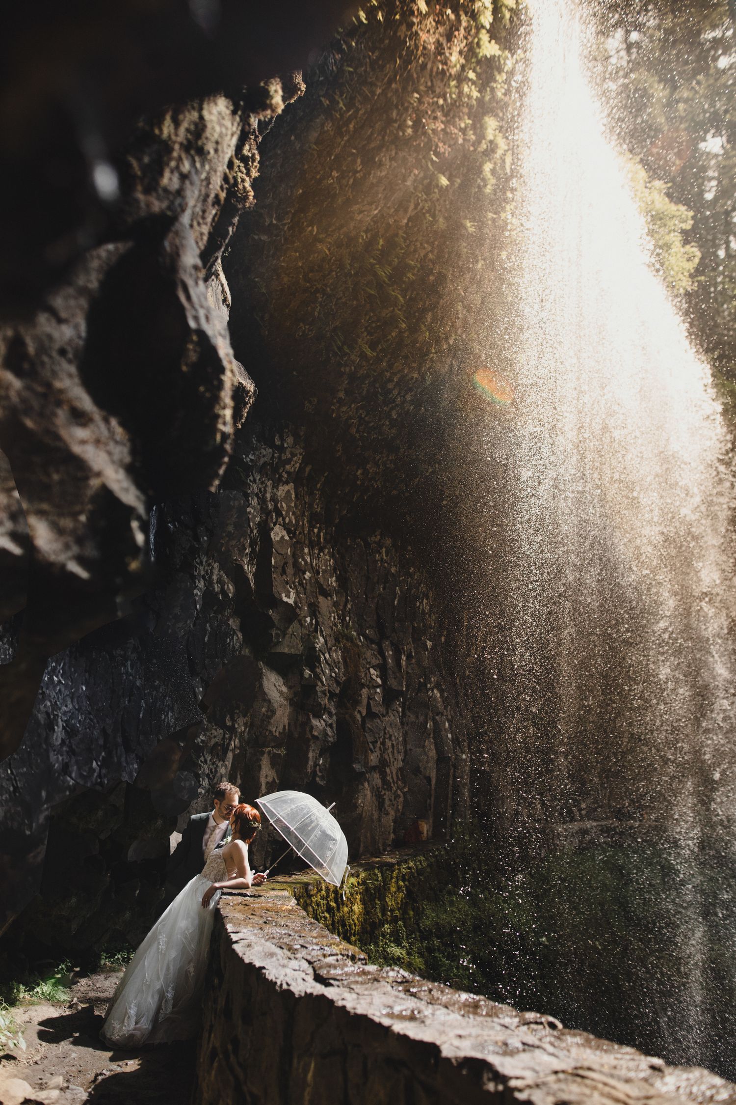 An elegant couple with an umbrella stands silhouetted against a dramatic ray of sunlight streaming into a rocky cave.