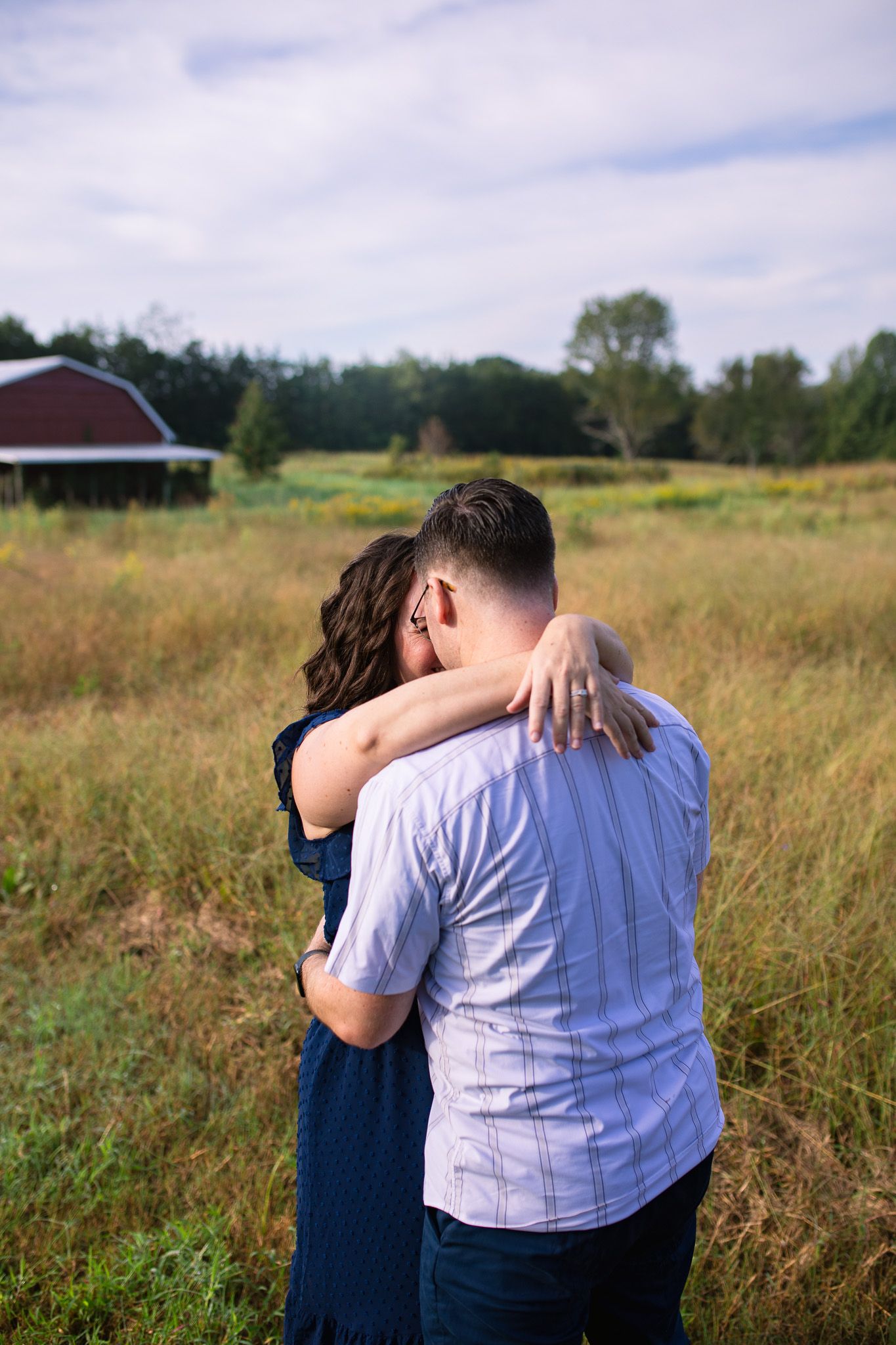 A couple embraces in a golden grass field with a red barn in the distance at Juniper Creek Farm