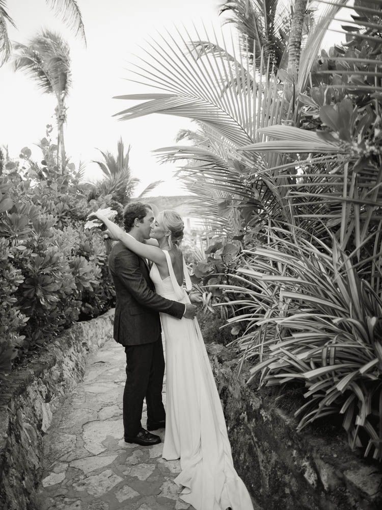A couple in formal wedding attire embrace in a harbor island, Bahama's coral sands hotel with palm trees and greenery surrounding them.